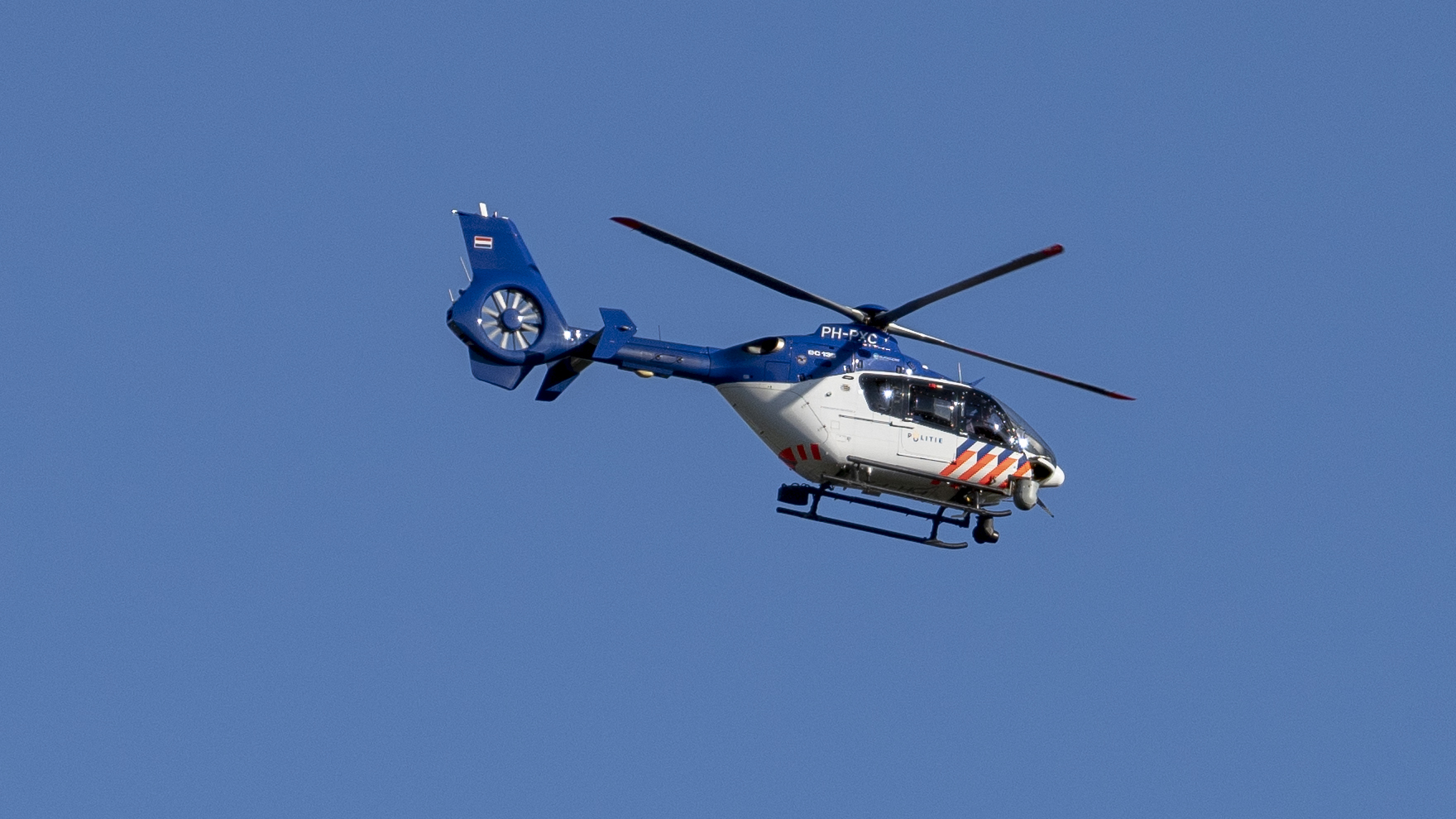 Een Nederlandse politiehelikopter in de lucht tegen een heldere blauwe hemel.