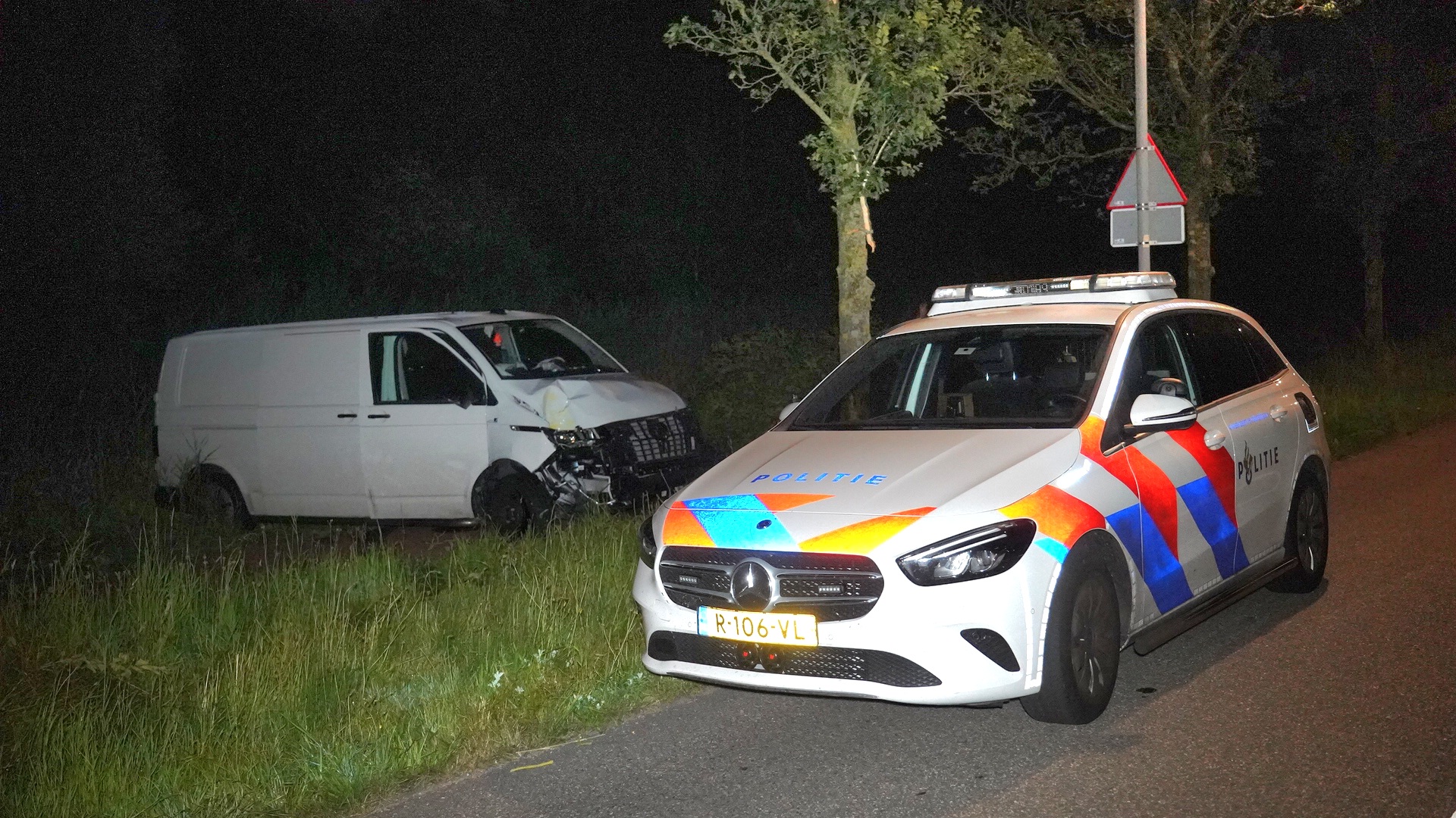 Witte bestelwagen met schade naast een politieauto op een donkere weg.