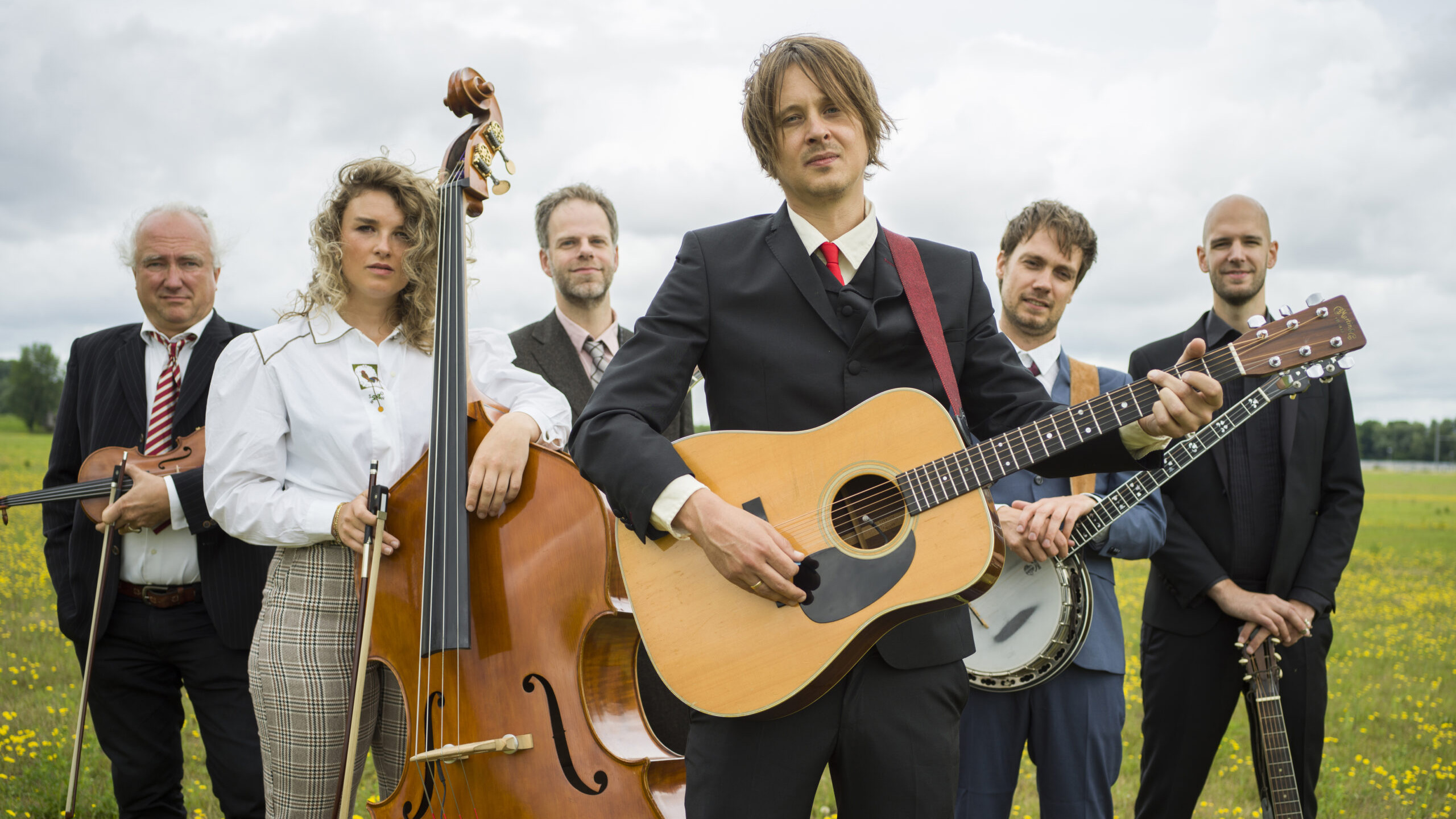 Een muziekgroep van zes personen in een veld met bloemen, waarbij verschillende instrumenten zoals een gitaar, banjo, contrabas en viool worden bespeeld.