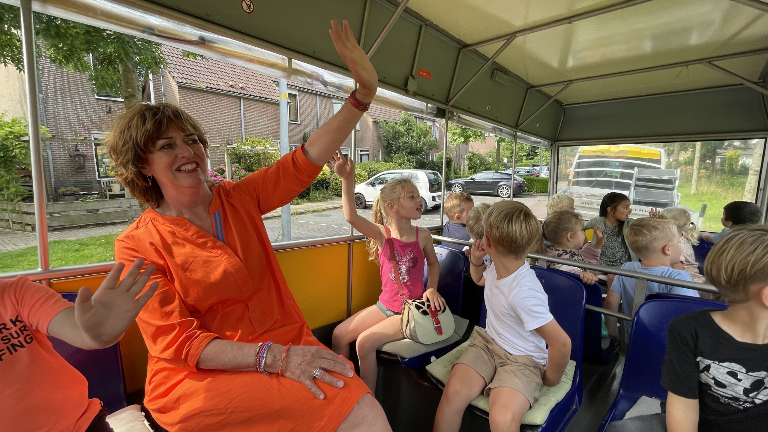 Een vrouw in een oranje jurk zit glimlachend in een bus omringd door kinderen.