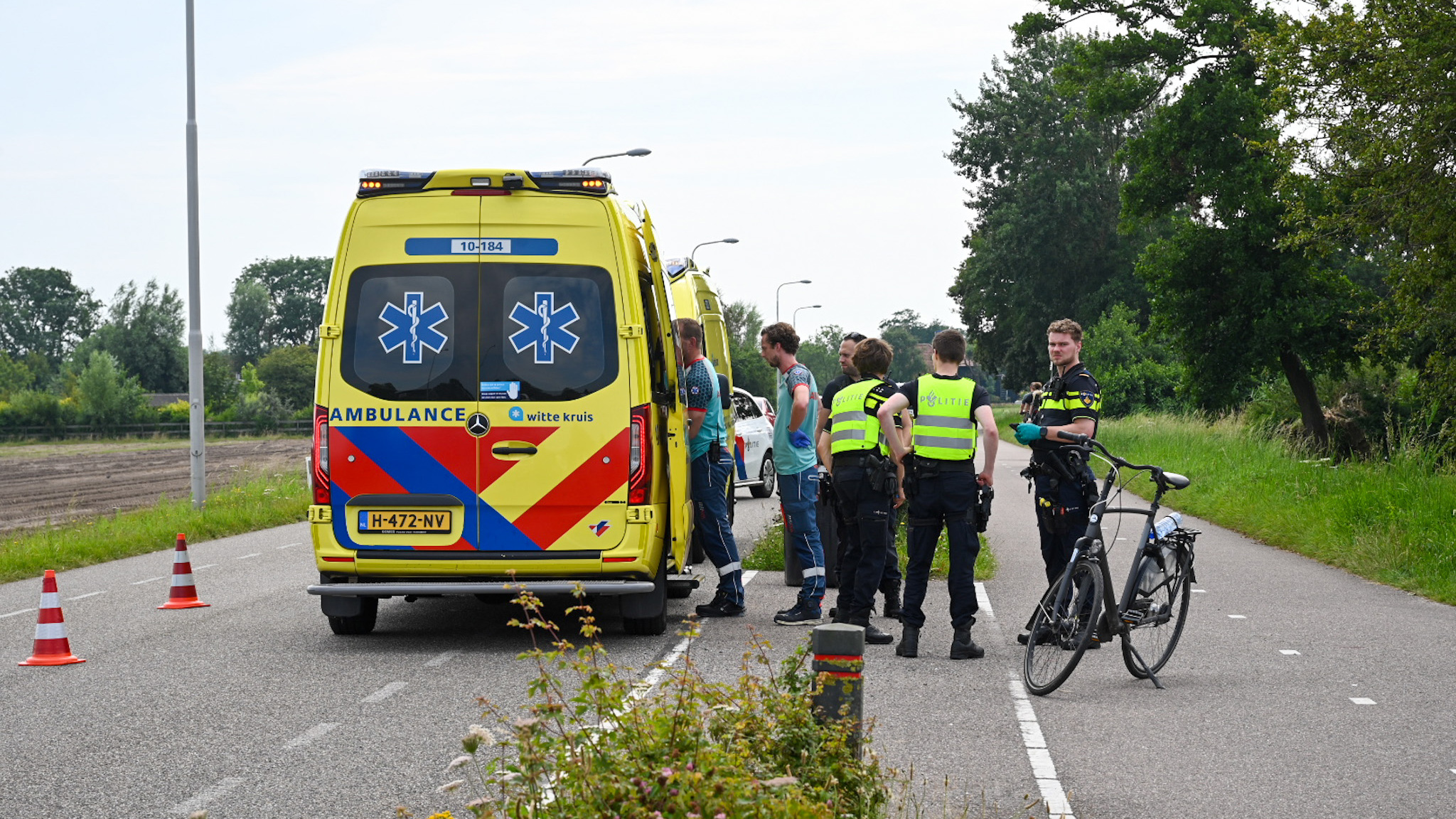 Politie en ambulancepersoneel bij ambulance op landweg, met fiets en verkeerskegels op de achtergrond.