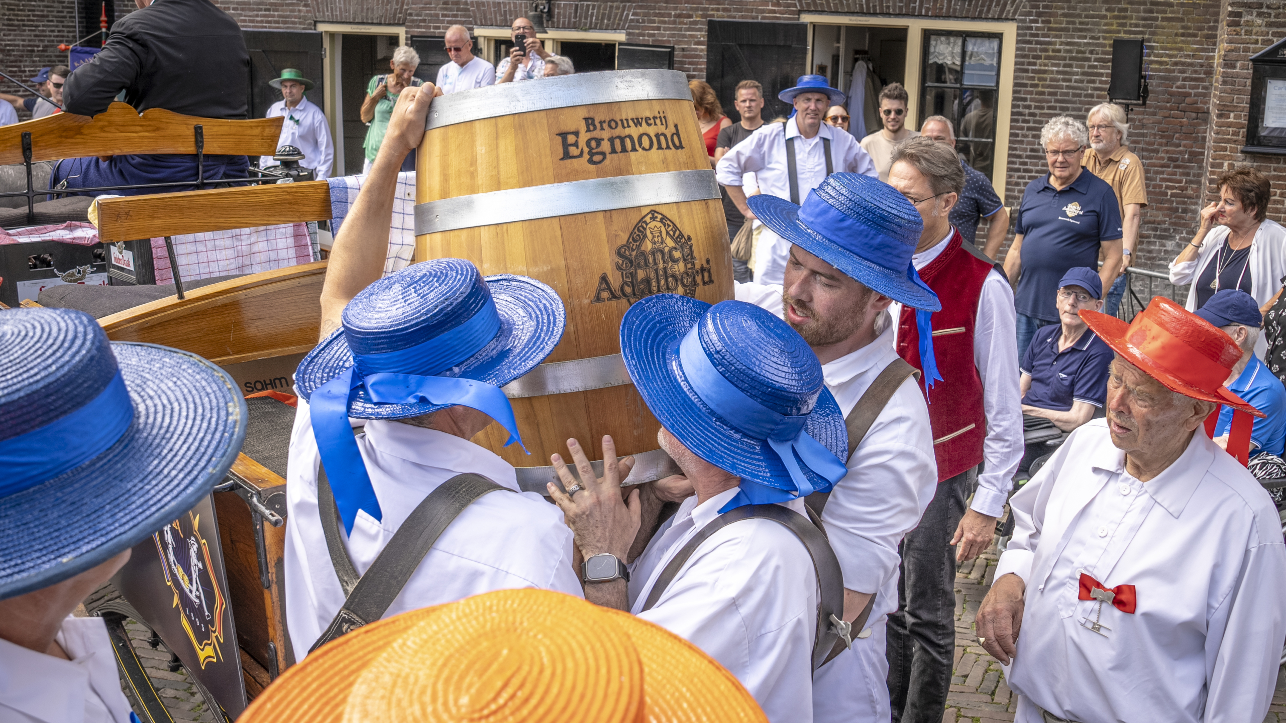 Mensen in kleurrijke hoeden tillen een biervat tijdens een festival of evenement, terwijl een groep toeschouwers toekijkt.