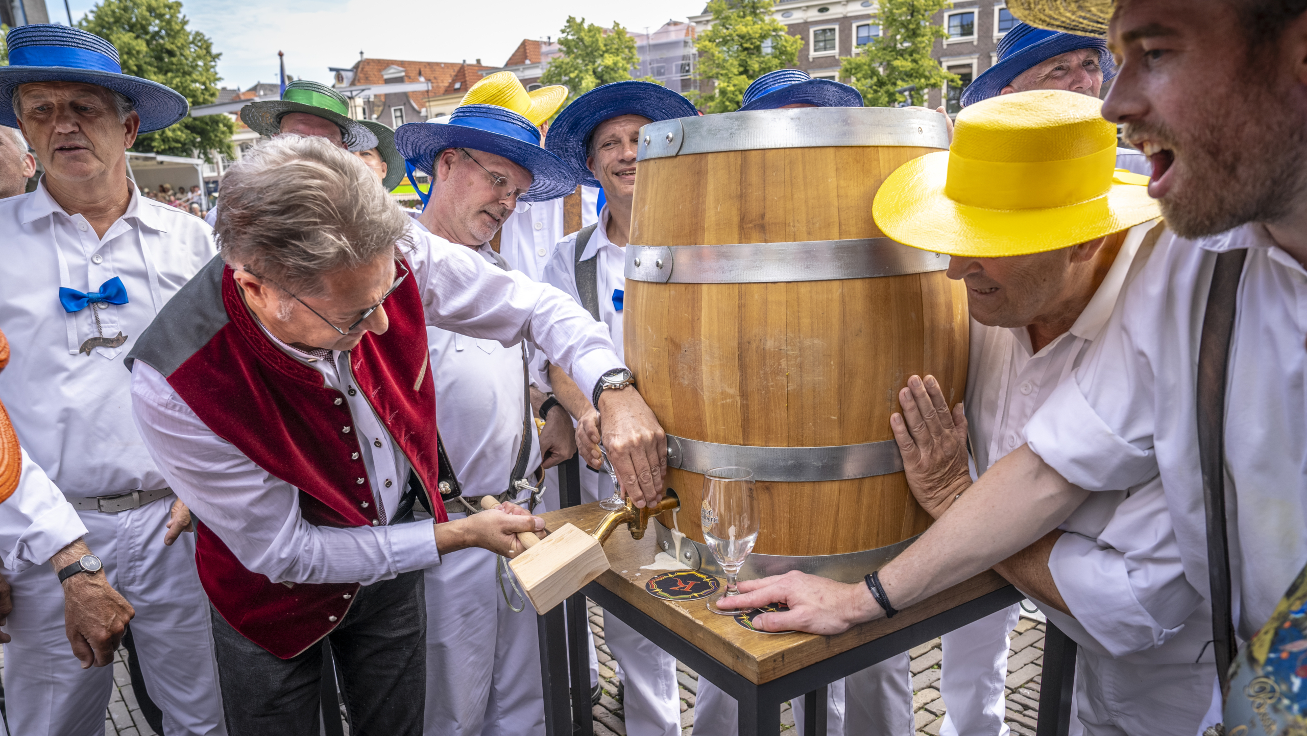 Mensen in traditionele kleding en kleurrijke hoeden tappen drank uit een houten vat op een straatfestival.