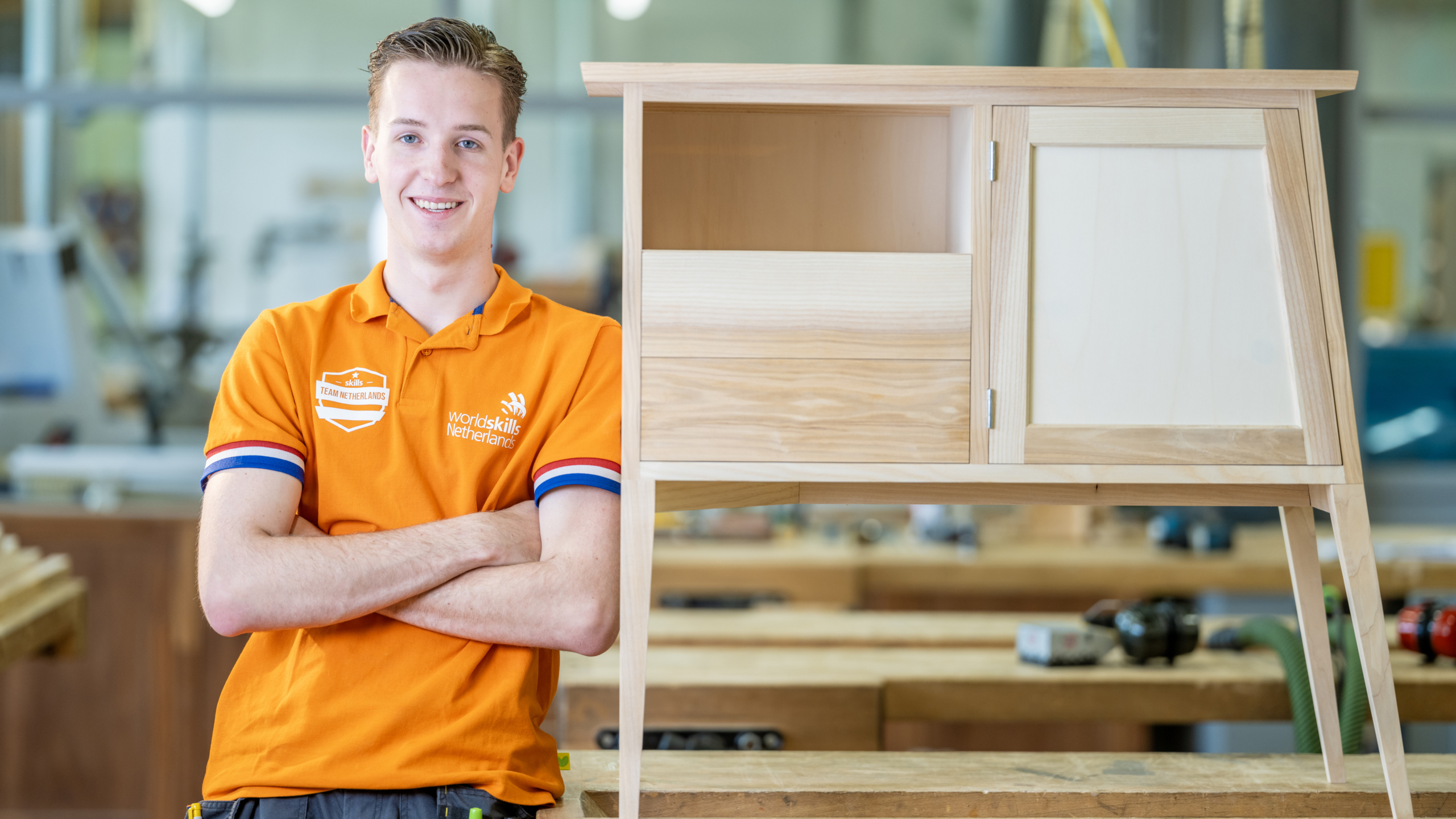 Man in oranje shirt van Team Netherlands bij een houten meubelstuk in werkplaats.