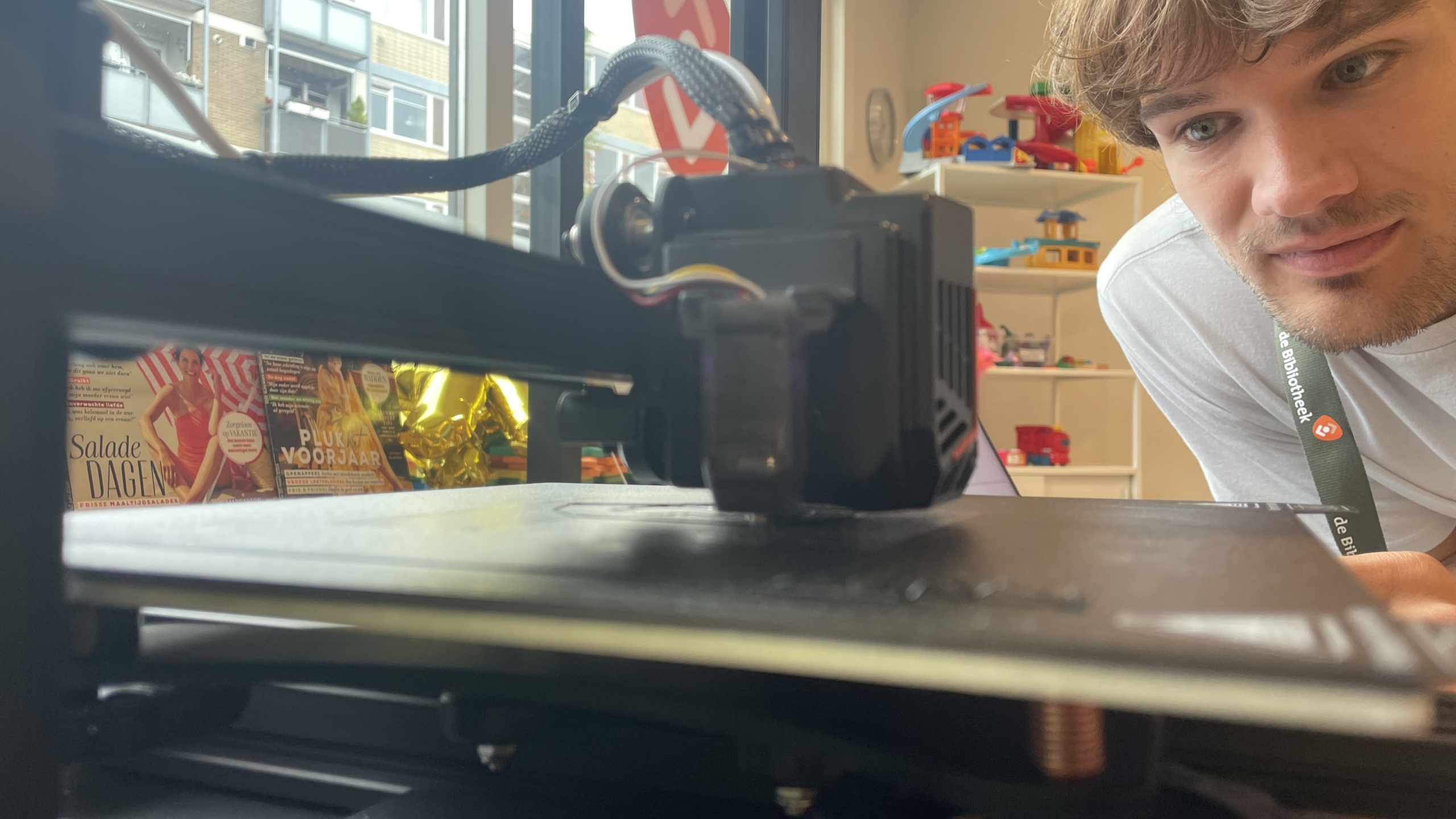 Man kijkt naar een 3D-printer in werking, met magazines en kinderspeelgoed op de achtergrond.