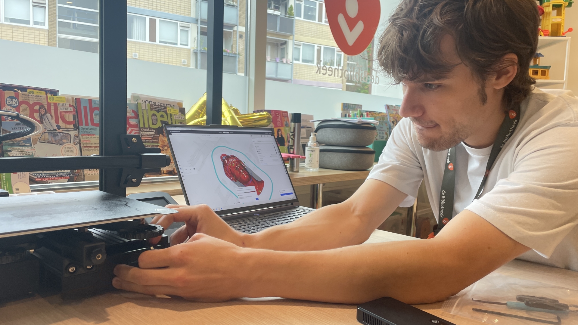 Man bedient een 3D-printer terwijl hij op een laptop een 3D-model bekijkt in een bibliotheekomgeving.