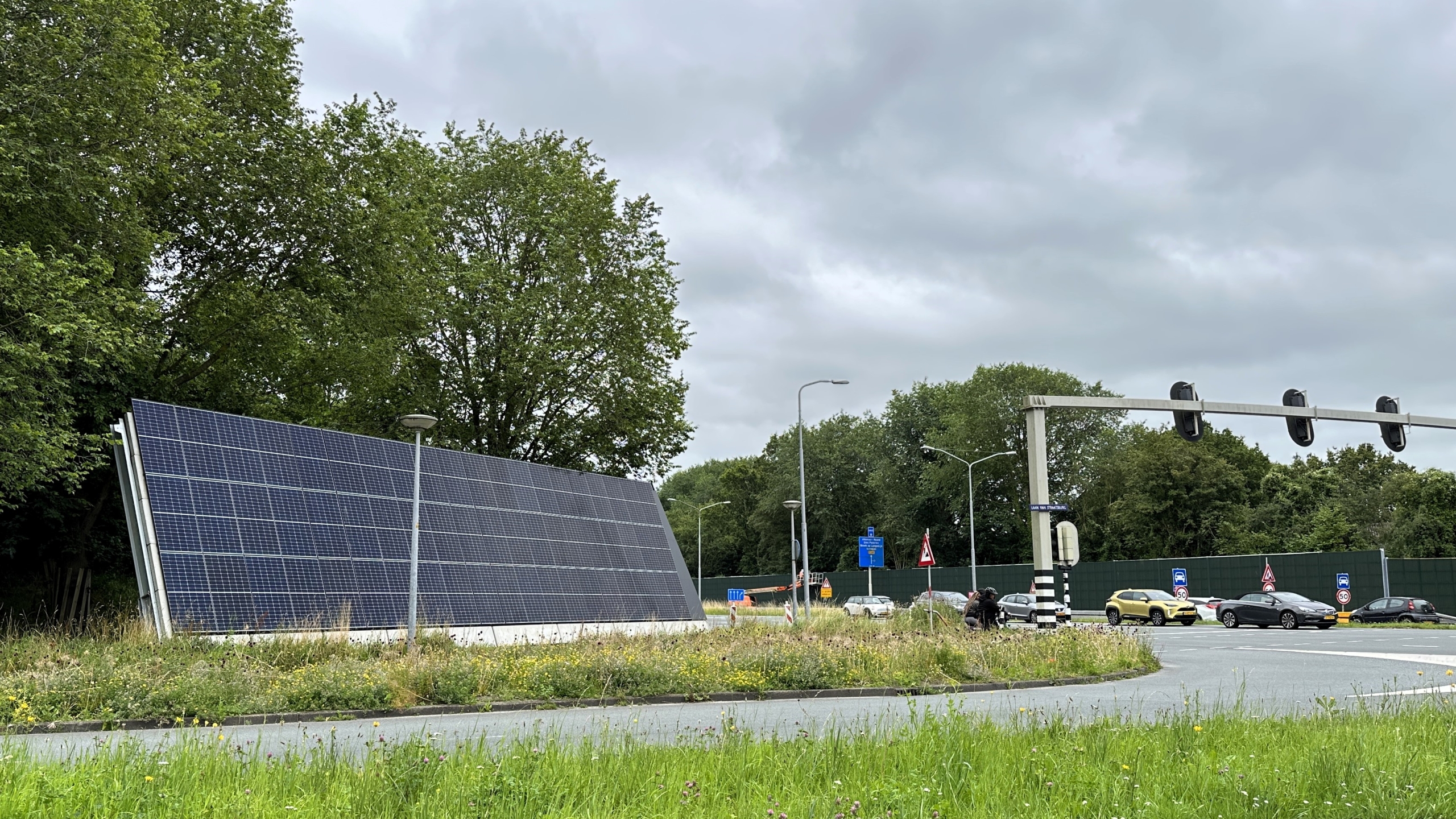 Zonnepanelen op een rotonde met verkeerslichten en auto's op de achtergrond, omgeven door bomen en gras.