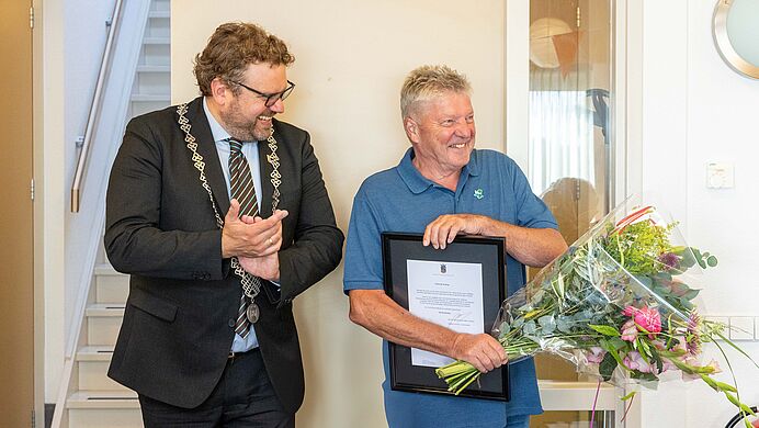 Twee mannen lachen; de man links draagt een keten en applaudisseert, terwijl de man rechts een ingelijste oorkonde en een boeket bloemen vasthoudt.