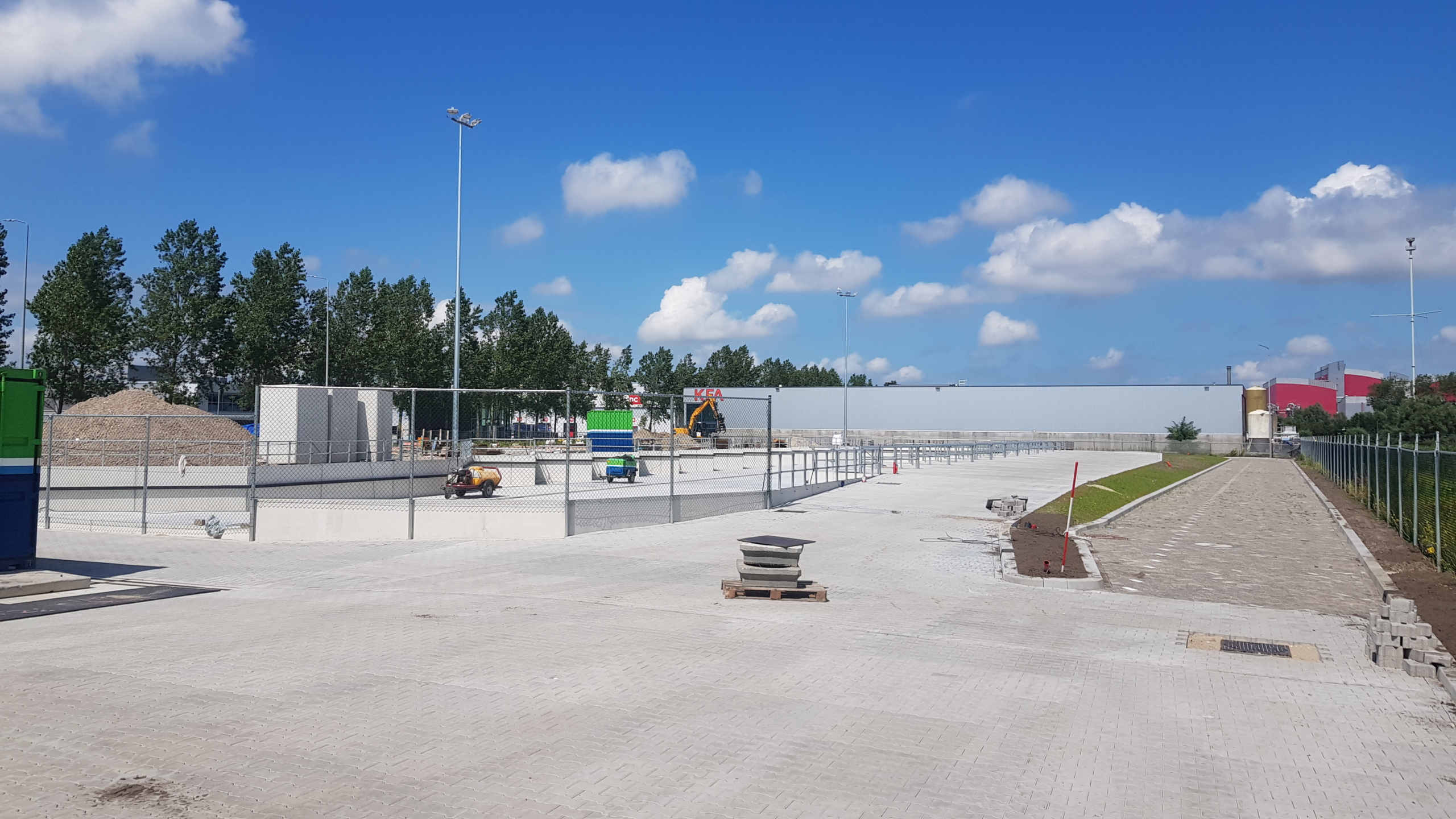 Betegeld terrein met hekwerk en bouwactiviteiten, op de achtergrond een bord met het opschrift "KEA" en bomenrij onder een blauwe lucht met wolken.