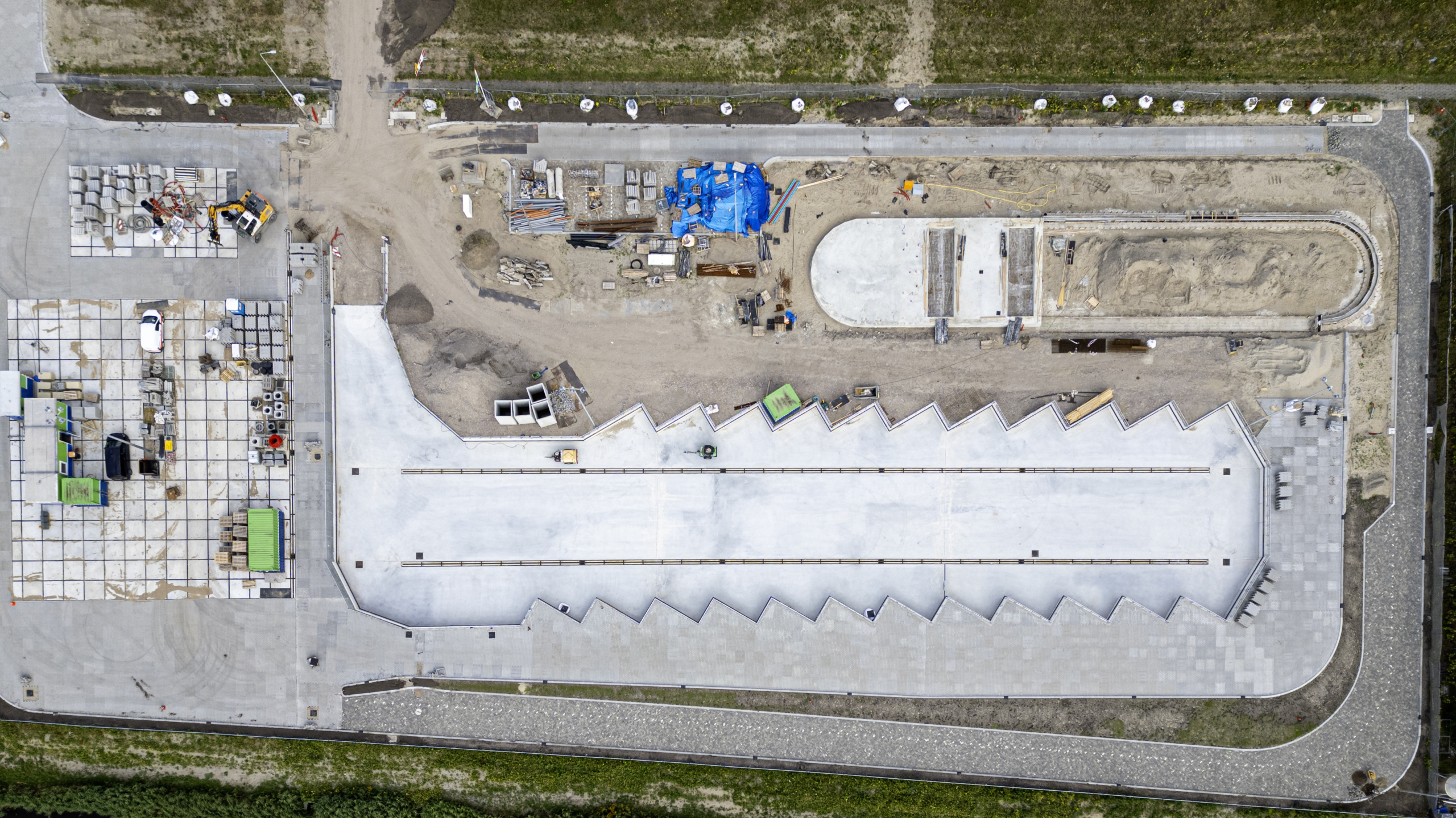 Luchtfoto van een bouwplaats met wegen, een rij zigzagvormige witte daken en bouwmaterialen verspreid over het terrein.