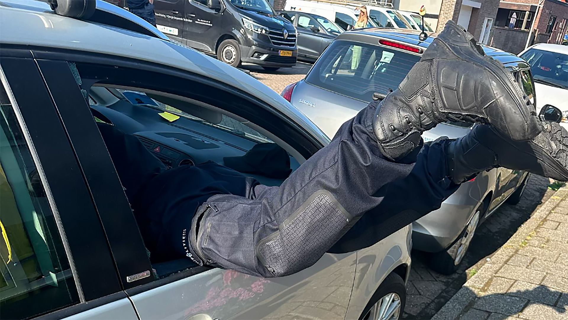 Persoon gekleed in uniform hangt met zijn benen uit een open autoraam op een parkeerplaats.