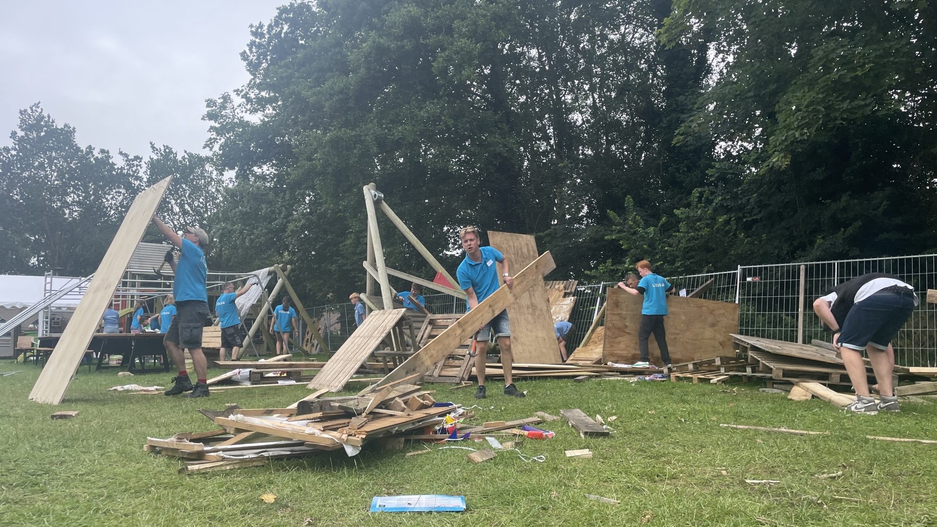 Mensen in blauwe shirts bouwen houten structuren op een grasveld.