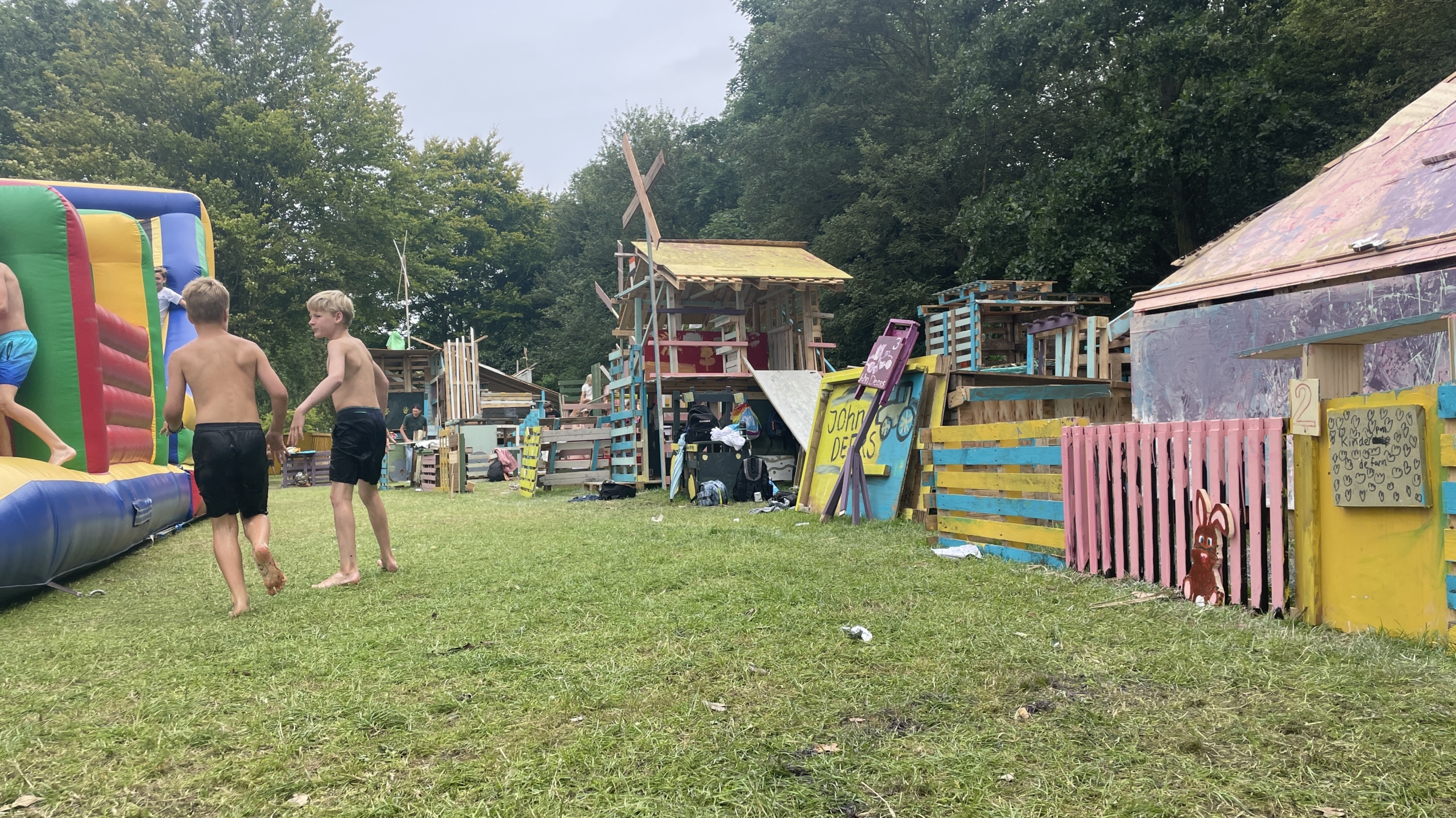 Kinderen spelen bij gekleurde houten hutten en een springkussen op een grasveld.