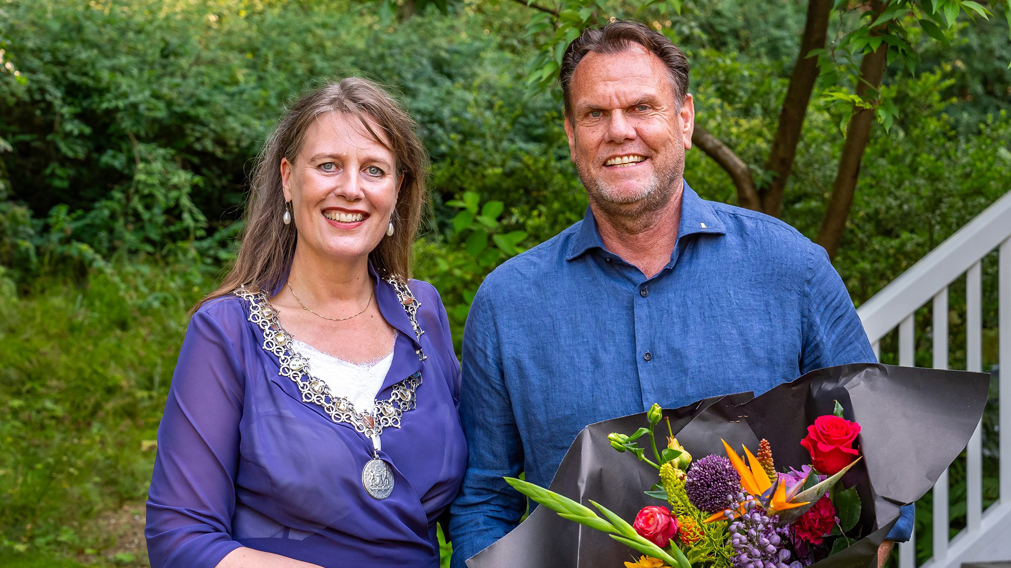 Een vrouw in een paarse jurk en een man in een blauwe blouse poseeren buiten met een boeket bloemen.