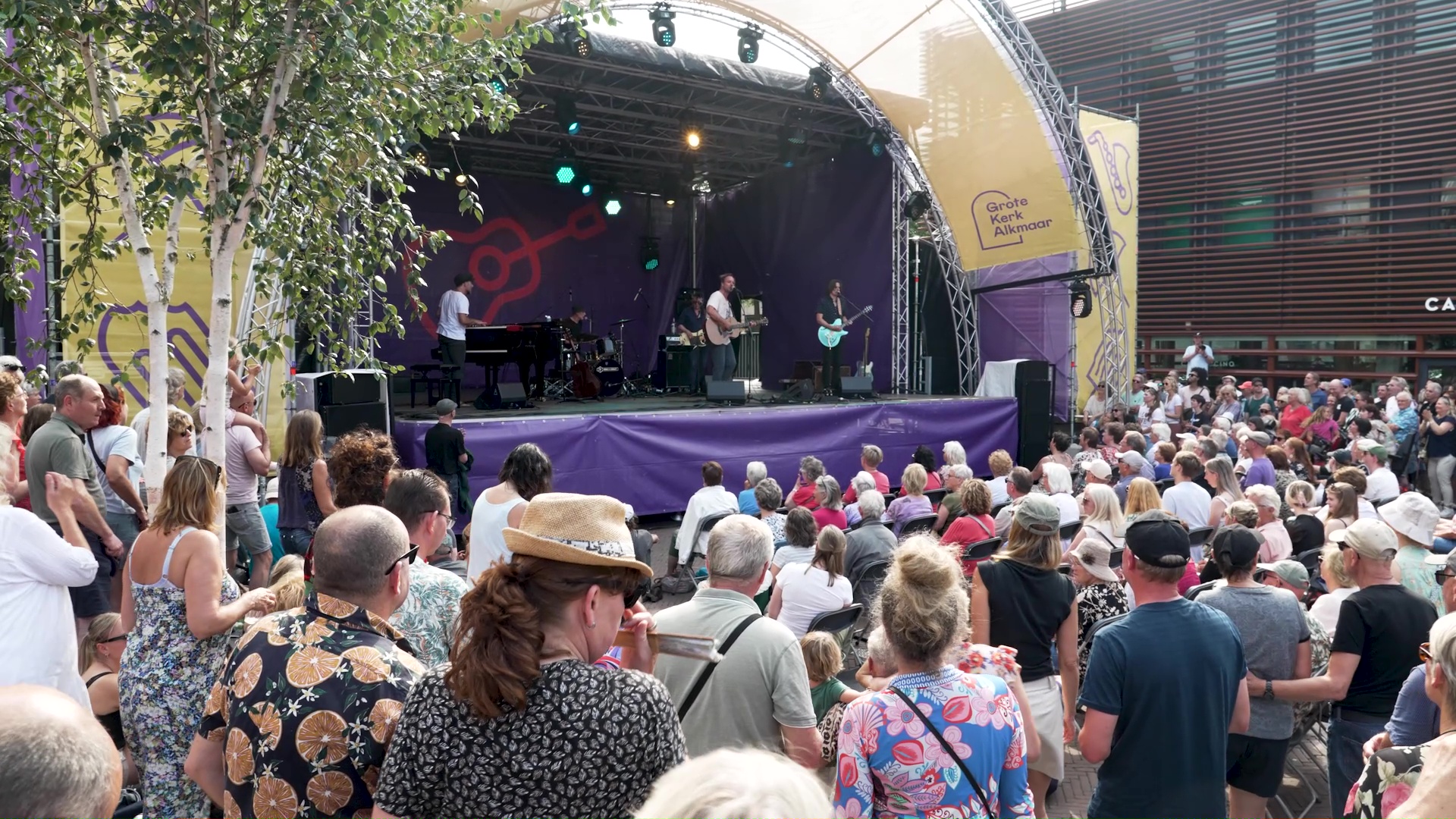 Een menigte mensen kijkt naar een live optreden op een buitenpodium bij Grote Kerk Alkmaar.