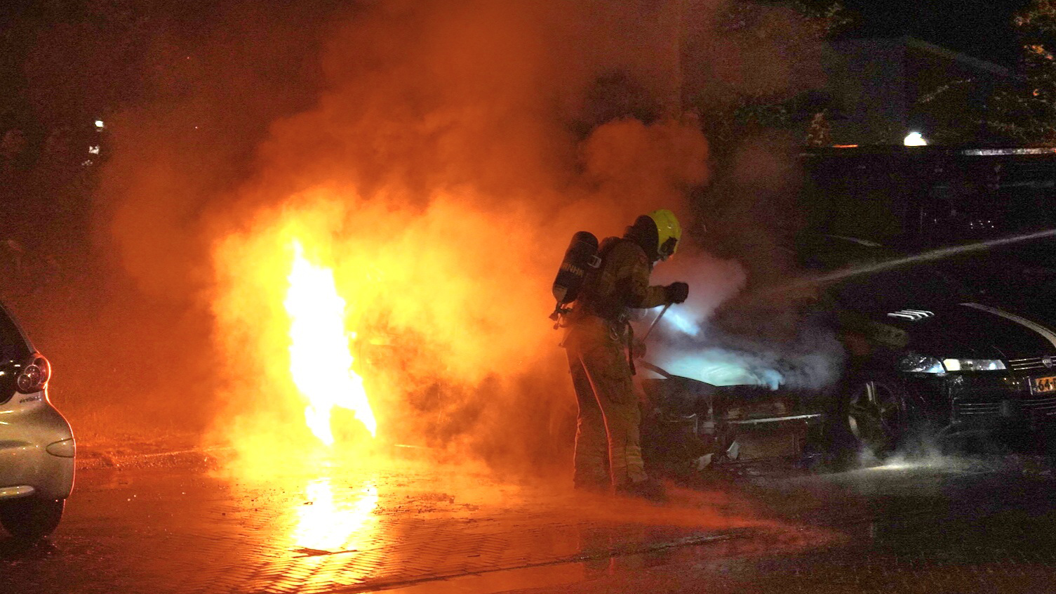 Brandweerman blust auto in brand in nachtelijke omgeving.