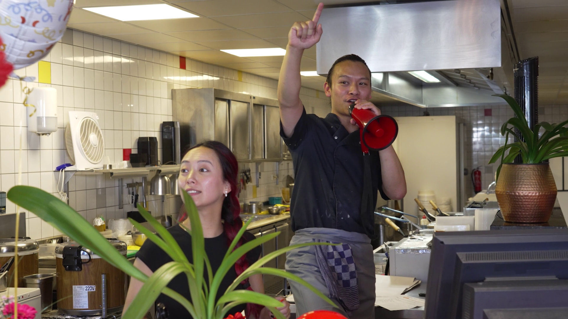 Twee mensen in een keuken; een man met een megafoon wijst omhoog en een vrouw kijkt in dezelfde richting.