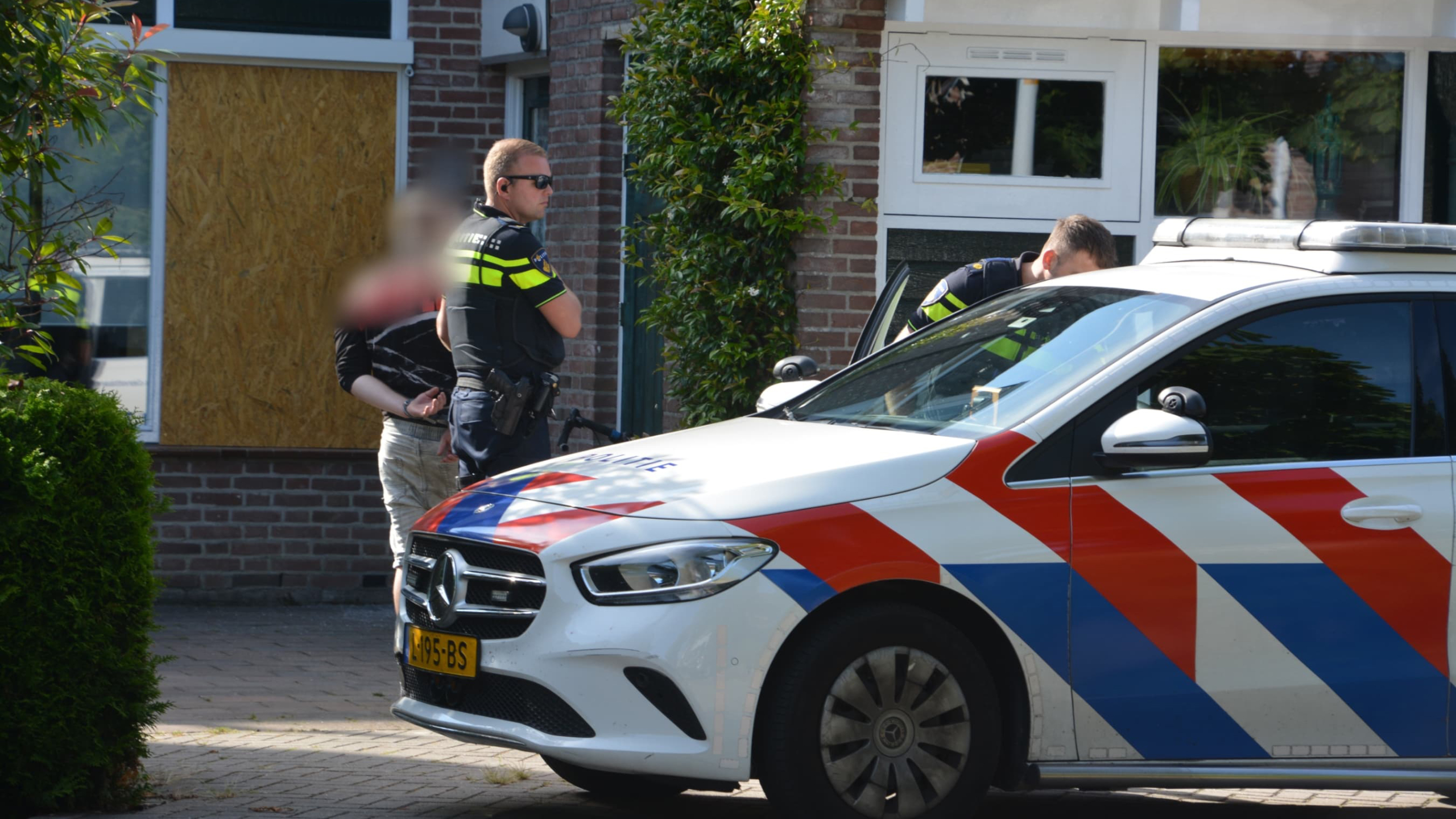 Twee politieagenten met een gearresteerde persoon naast een politieauto buiten een gebouw.