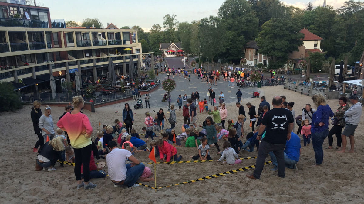 Mensen verzamelen zich op een zandheuvel bij een plein met winkels en restaurants, omgeven door groen. Kinderen spelen in het zand, terwijl volwassenen toekijken en praten.