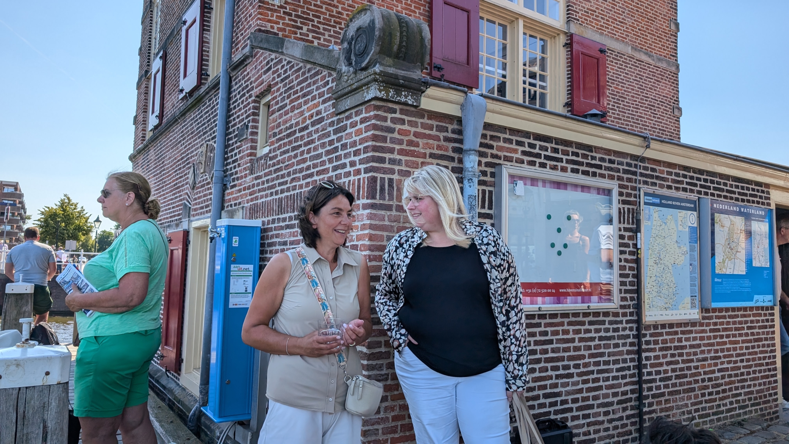 Twee vrouwen praten naast een bakstenen gebouw met informatieborden en een blauwe paal.