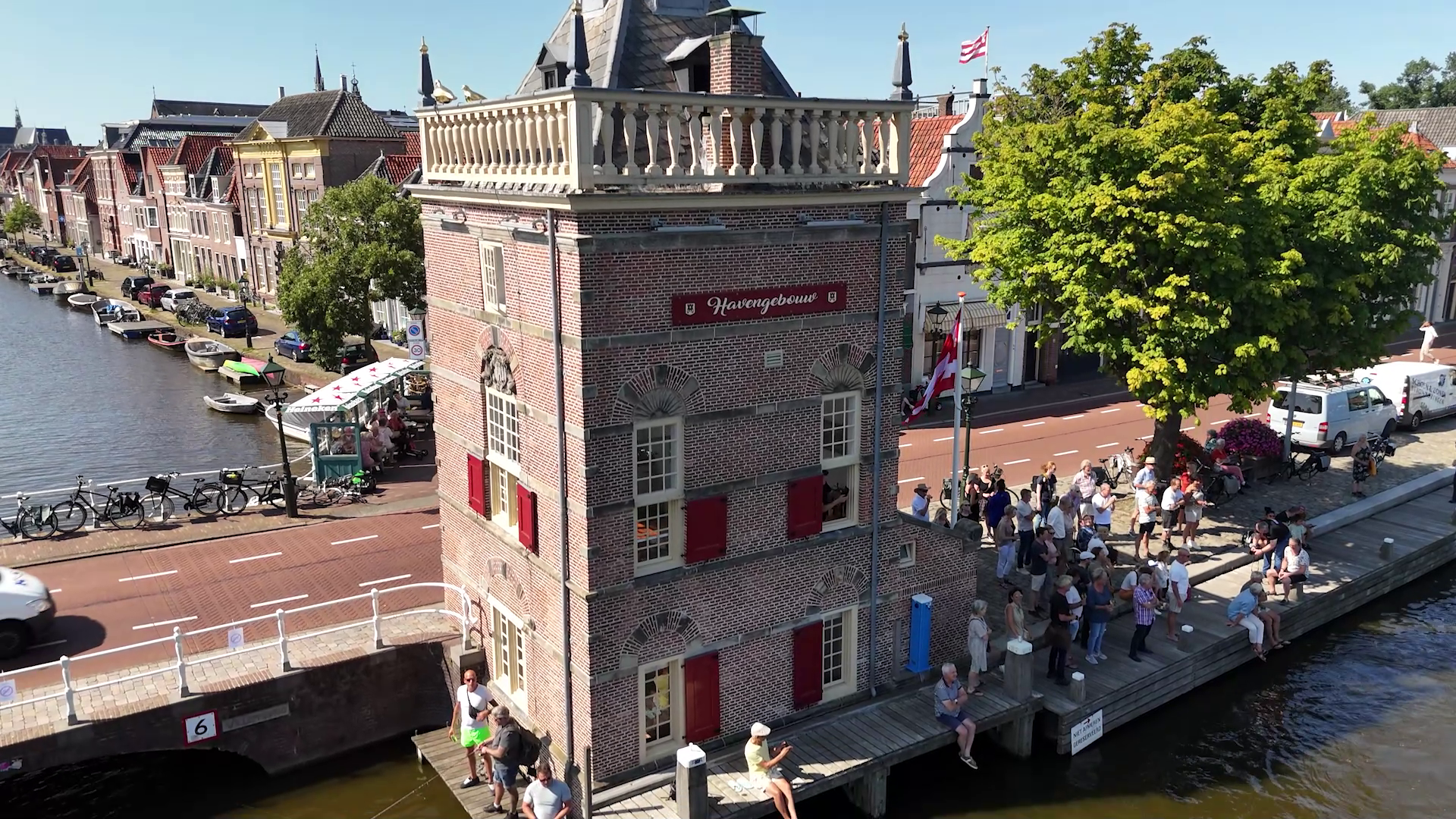 Havenggebouw bij gracht met fietsen, mensen en boten, zonnige dag in Nederlandse stad.