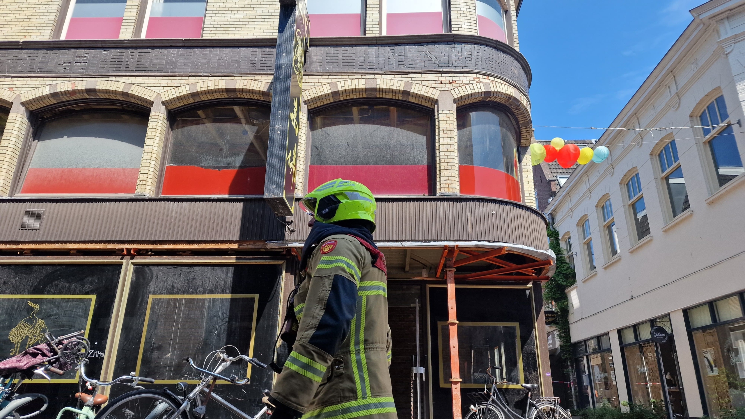 Brandweerman in uniform met fluorgele helm staat voor een bakstenen gebouw met ronde ramen, naast een straat vol fietsen en kleurrijke ballonnen aan een draad.