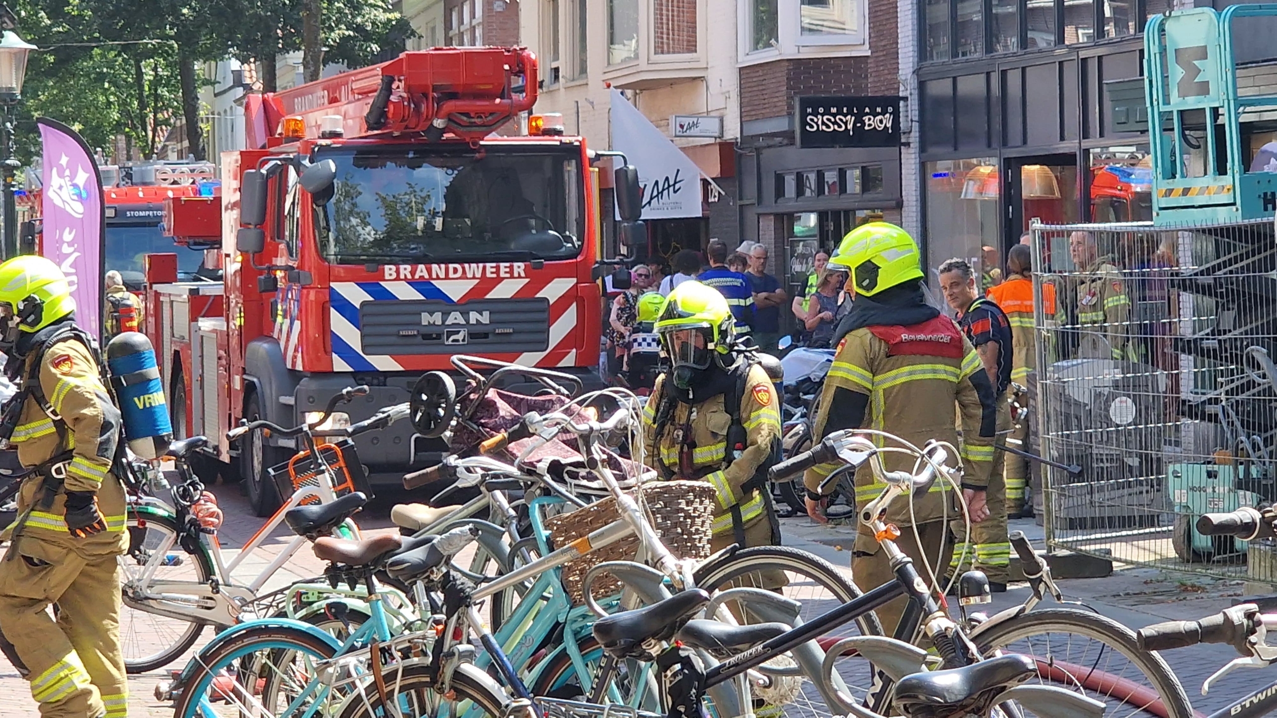 Brandweerlieden in uniform werken op een drukke straat met geparkeerde fietsen en brandweerwagens op de achtergrond.