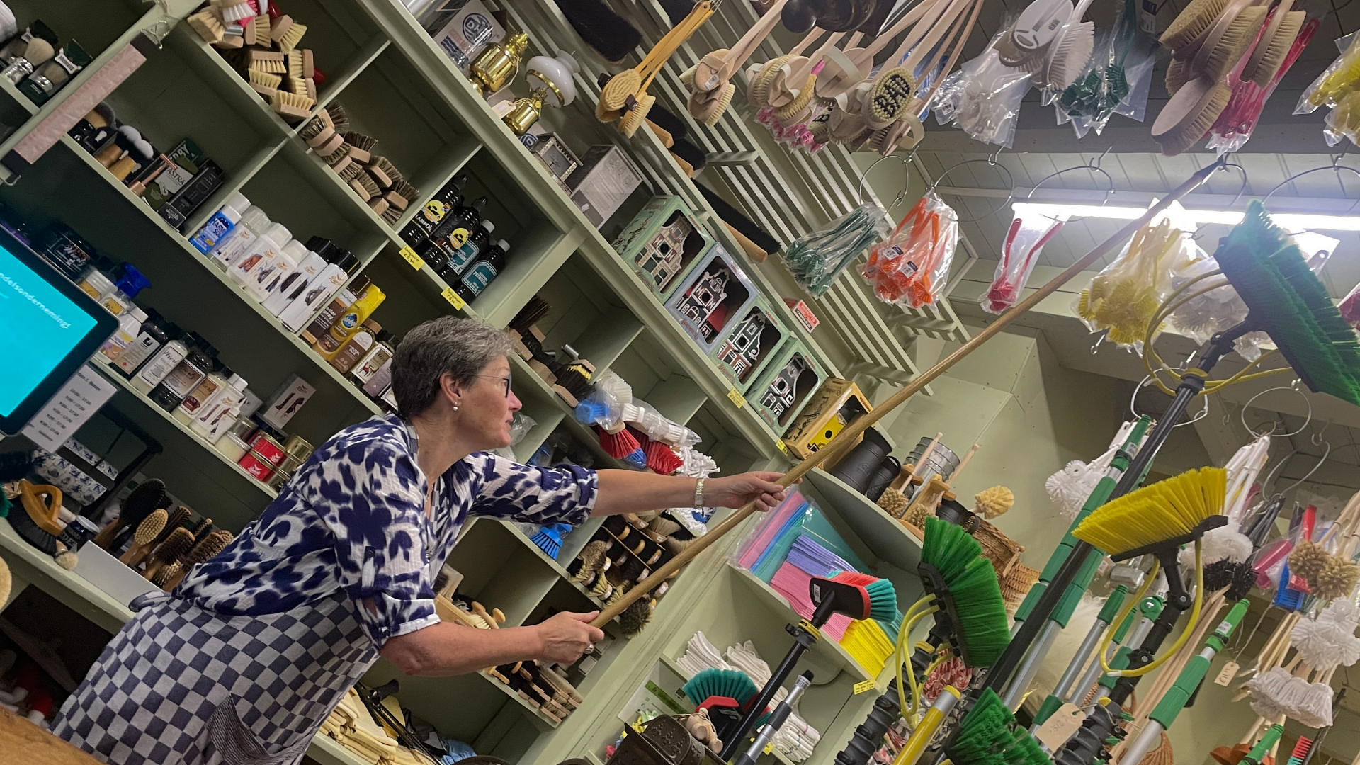 Een vrouw in een huishoudwinkel reikt naar borstels die aan het plafond hangen. De winkel heeft veel rekken met schoonmaakartikelen en huishoudelijke benodigdheden.