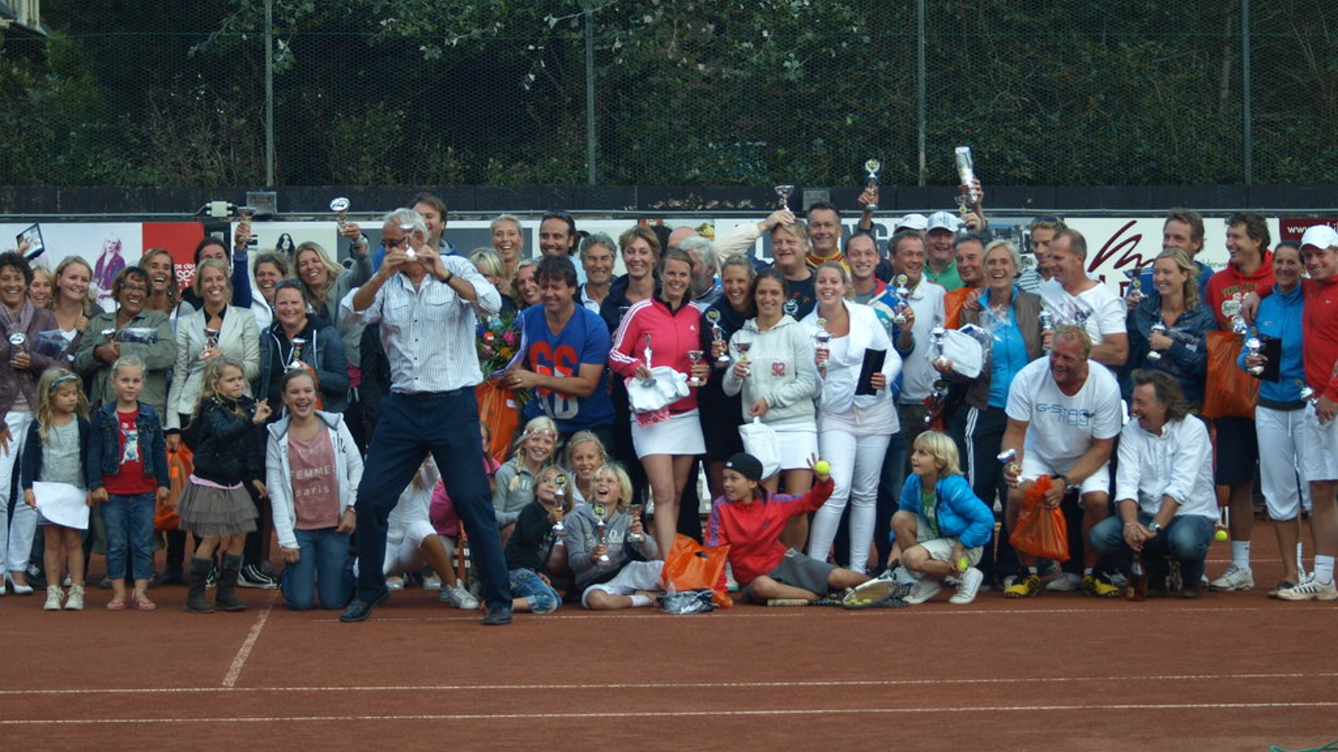 Grote groep mensen poseert met bekers op een tennisbaan.