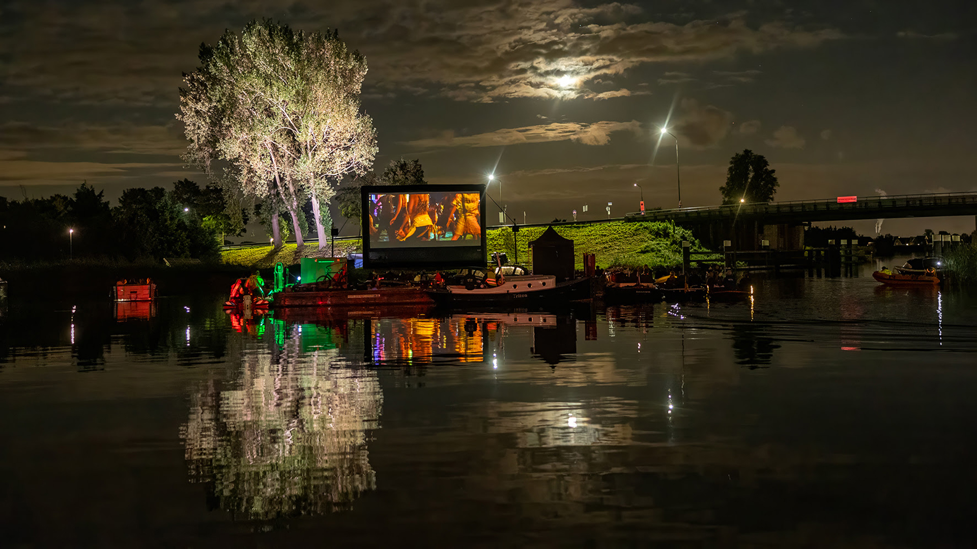 Bioscoopscherm op een rivier met boten tijdens een nachtelijke filmvoorstelling, verlicht door straatlantaarns en maanlicht.