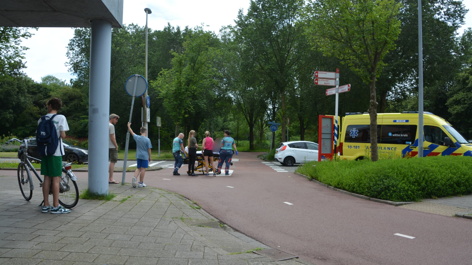 Mensen staan rond een ambulance op een fietspad, terwijl paramedici een brancard voortduwen.