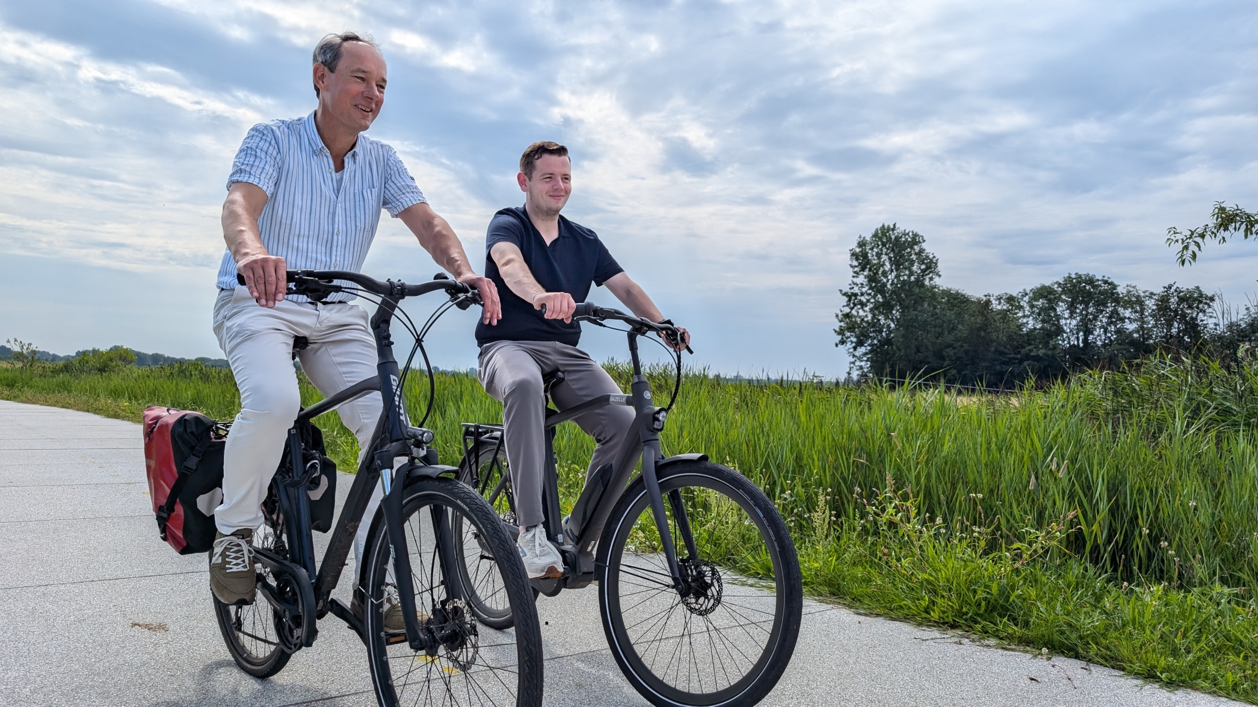 Twee mannen fietsen over een pad door een groen landschap met gras en bomen.