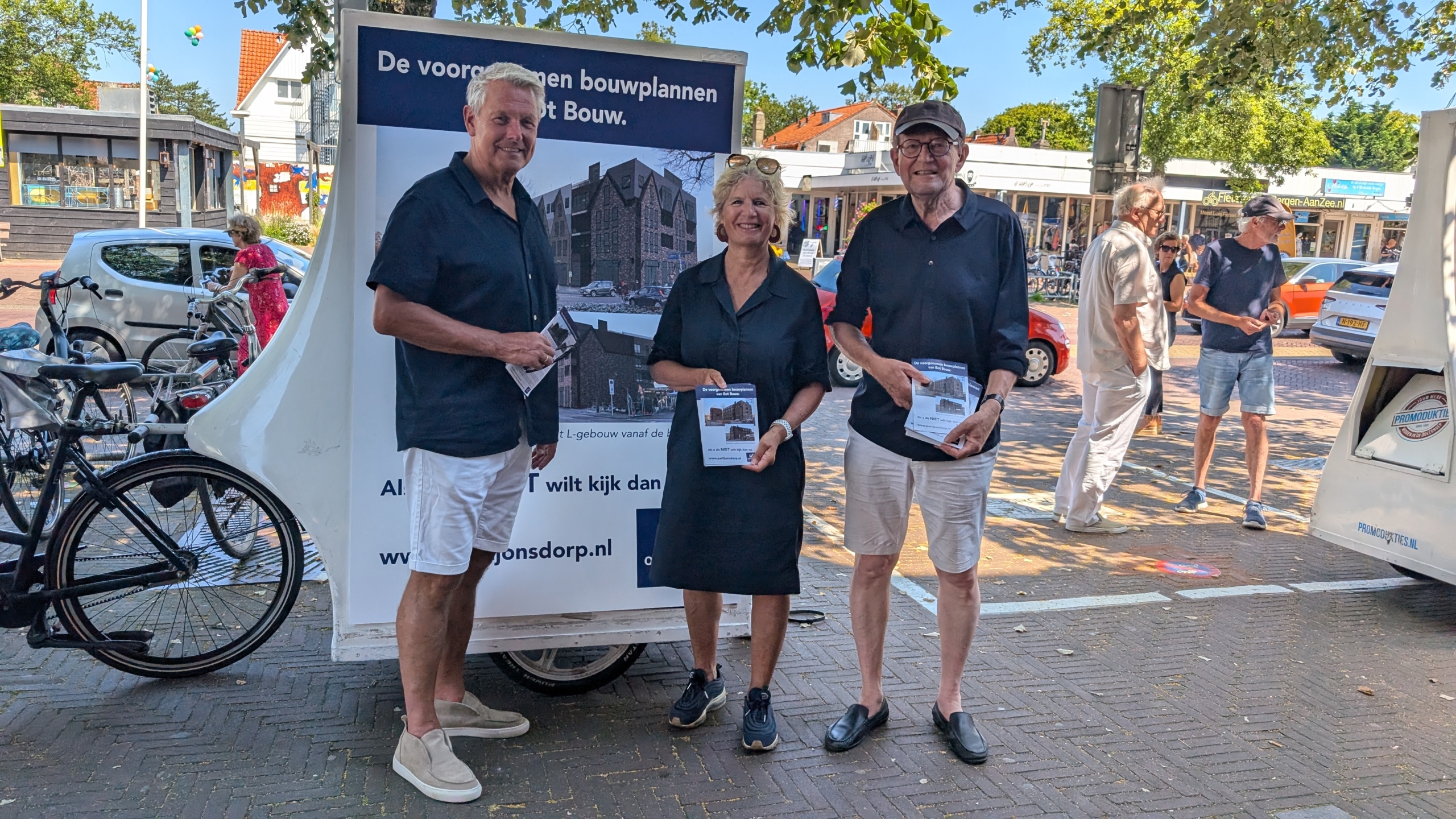 Drie mensen poseren voor een bord met bouwplannen op straat, terwijl ze brochures vasthouden.