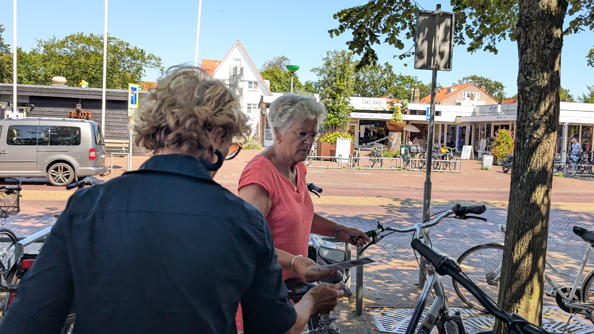 Twee vrouwen met fietsen op een plein, met winkels en een parkeerterrein op de achtergrond.