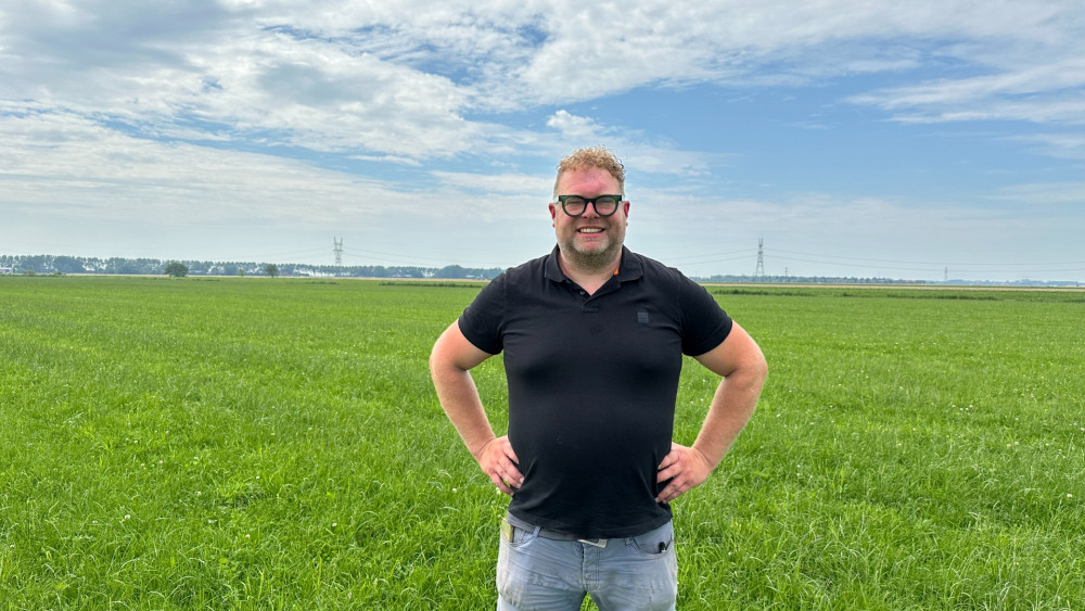 Man in een zwart shirt staat met zijn handen op zijn heupen in een groen weiland onder een bewolkte hemel.