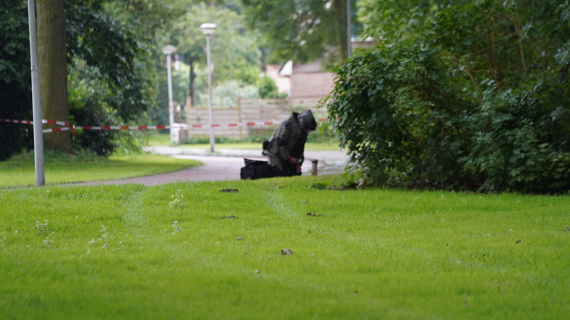 Man in beschermingspak werkt op een grasveld, rood-wit afzetlint op de achtergrond.