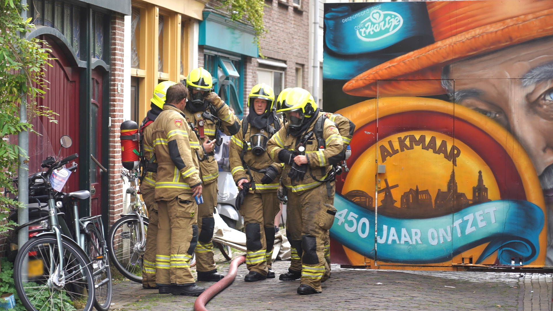 Brandweerlieden in uitrusting staan voor een deur in een straat tegen een kleurrijke muurschildering met de tekst "ALKMAAR 450 JAAR ONTZET".