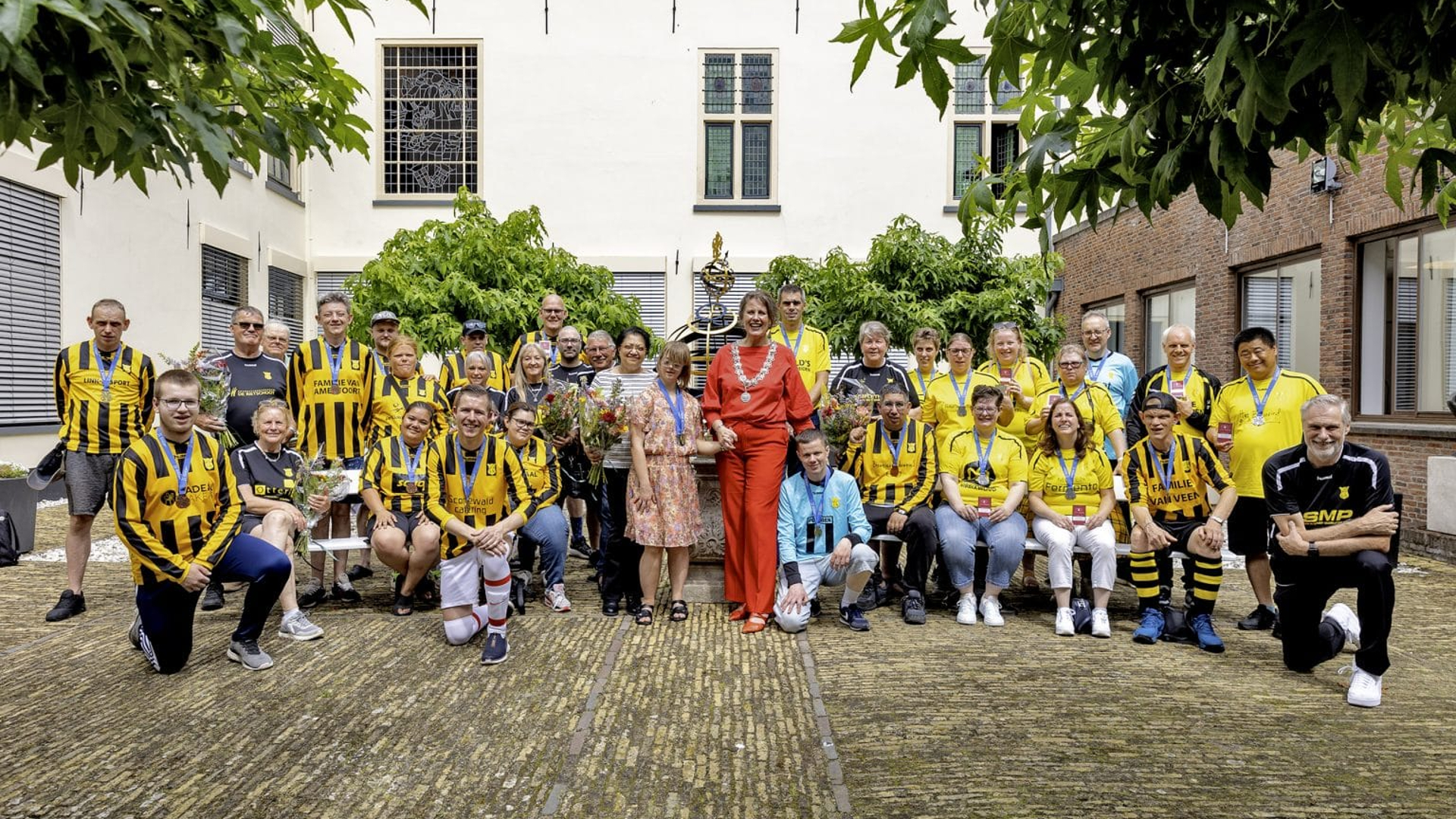 Groepsfoto van een voetbalteam in gele en zwarte tenues, samen met een vrouw in een rood pak met een ambtsketen, in een binnentuin.