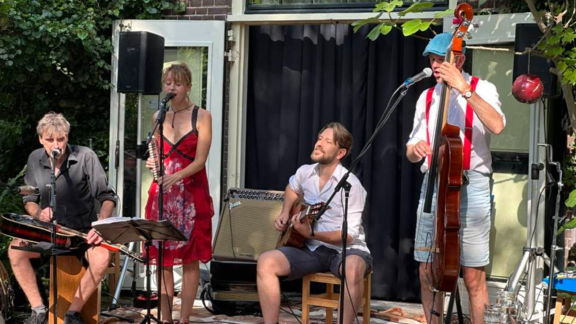 Muziekband met vier leden, inclusief zangeres met melodica, gitarist, bassist en percussionist, optreden buiten bij een huis.