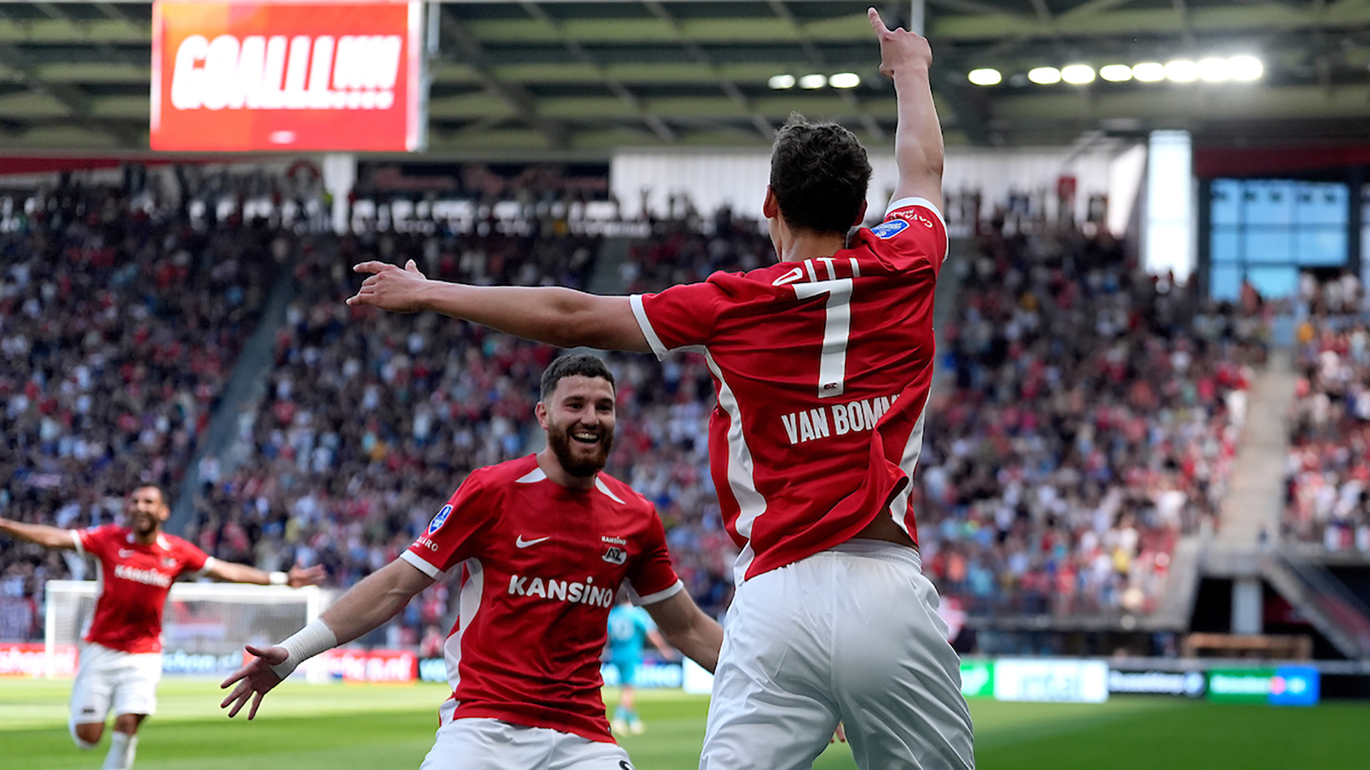 Voetballers in rood-witte tenues vieren een doelpunt in een vol stadion.