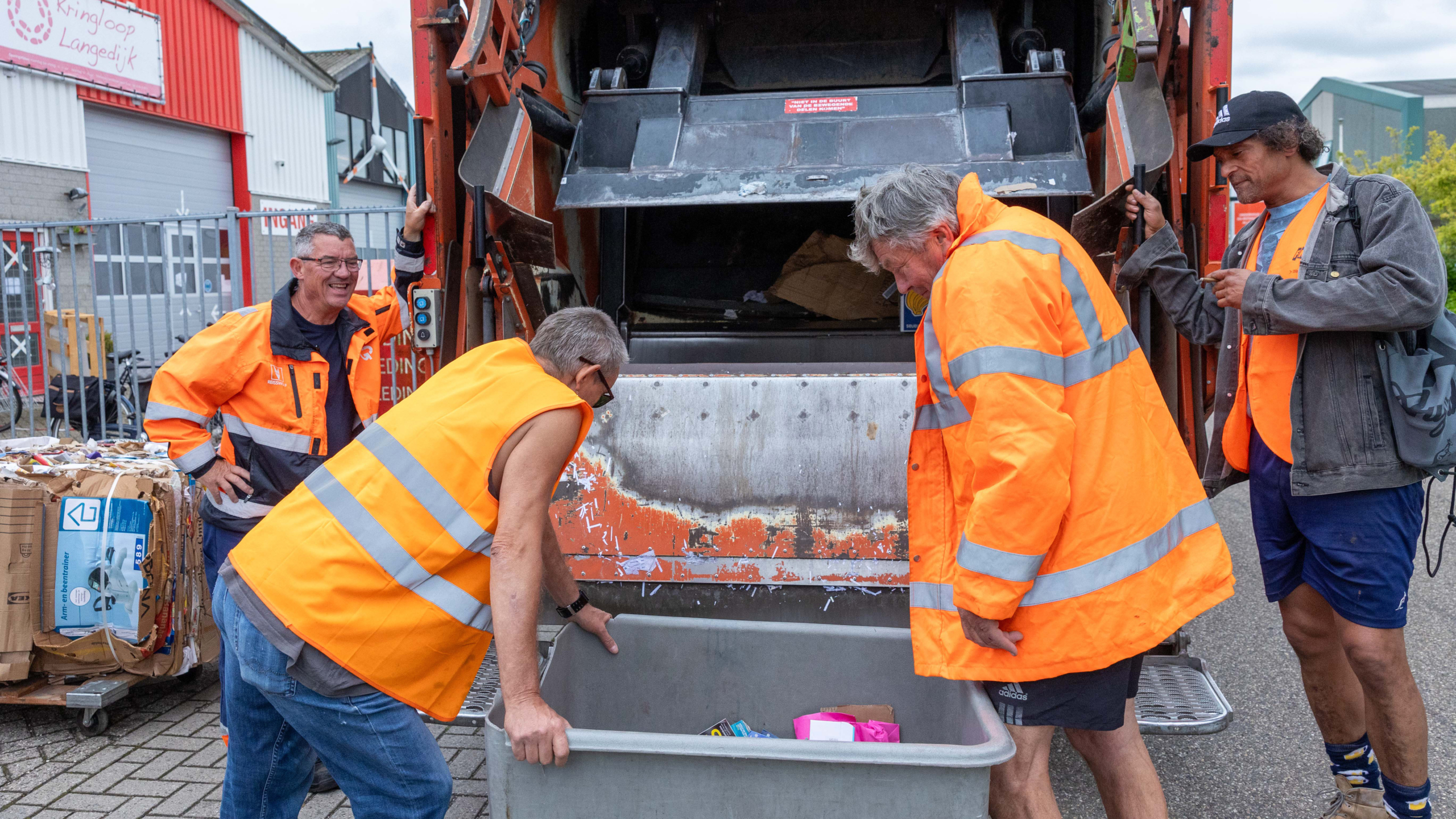 Vuilnismannen in feloranje veiligheidsvesten werken samen bij een vuilniswagen naast een kringloopwinkel.