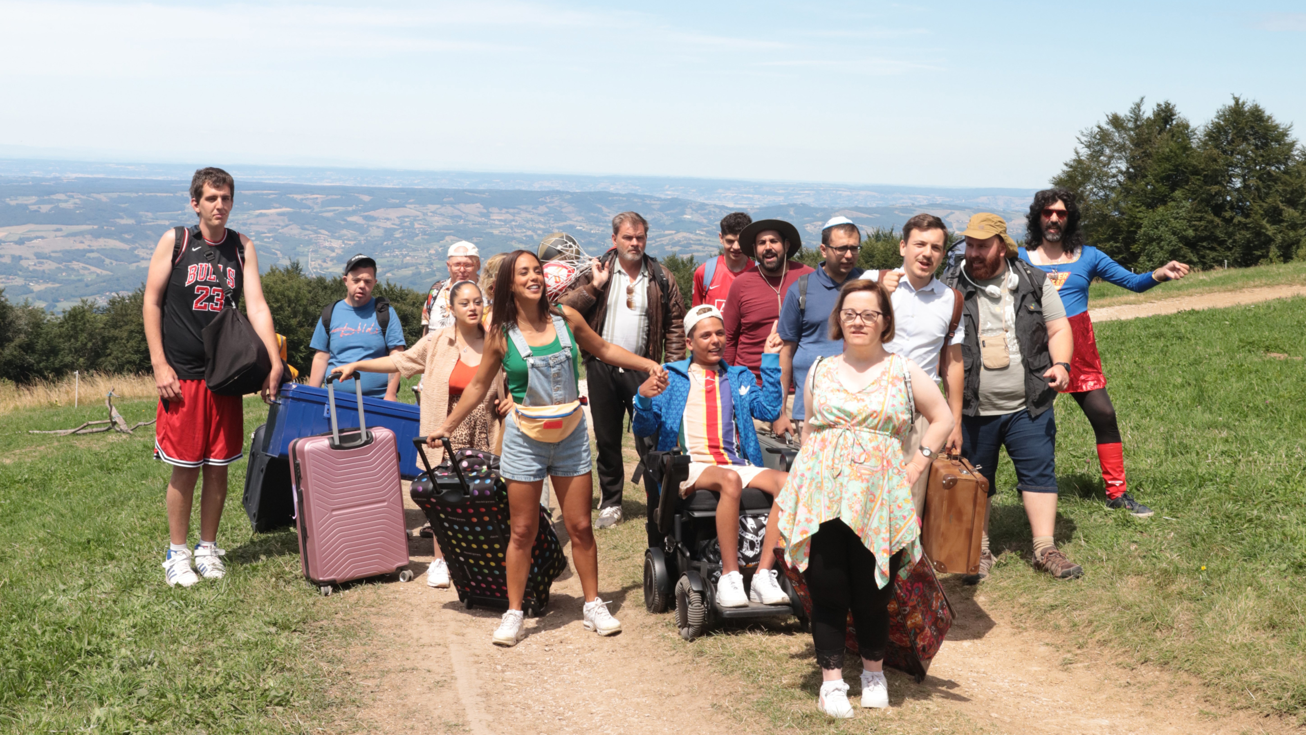 Een diverse groep mensen poseert buiten op een wandelpad met bagage, tegen een achtergrond van heuvels en een ver uitzicht.