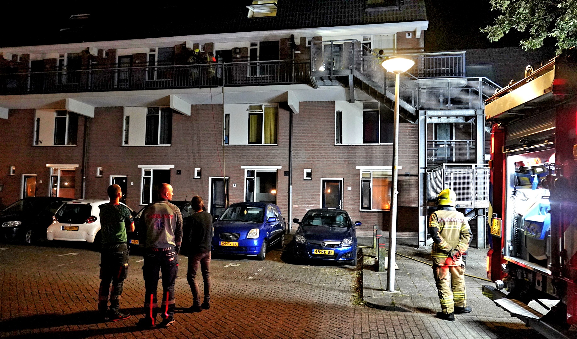 Brandweerlieden en omstanders voor een appartementencomplex, waar een brandweerauto geparkeerd staat bij een nachtelijke noodsituatie.