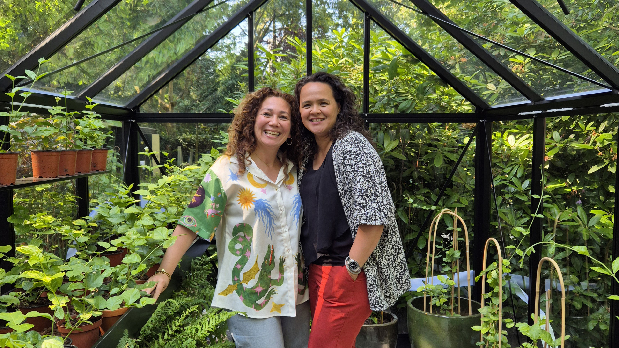 Twee vrouwen lachen in een kas vol met groene planten.