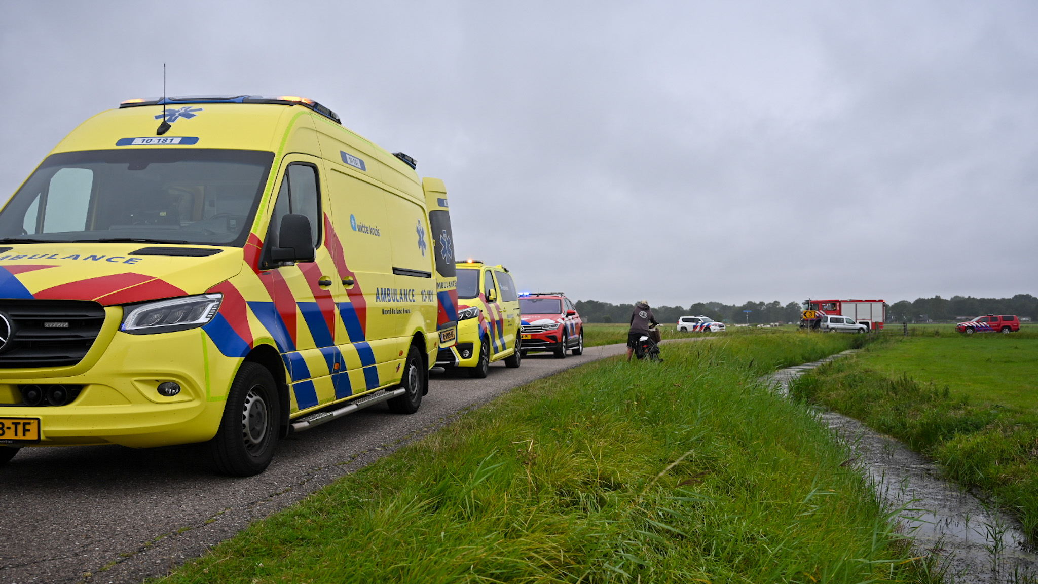 Rij ambulances en noodvoertuigen langs een weg in een landelijke omgeving op een bewolkte dag.