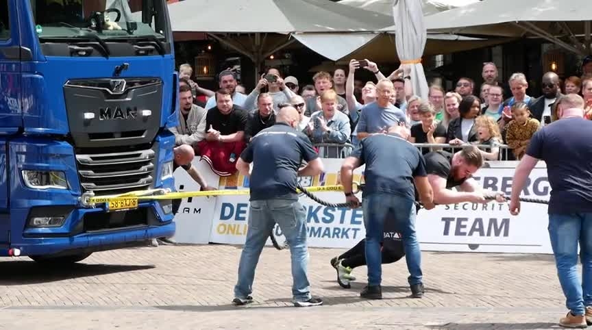 Man probeert een vrachtwagen te trekken, bijgestaan door meerdere mensen, terwijl een menigte toekijkt.