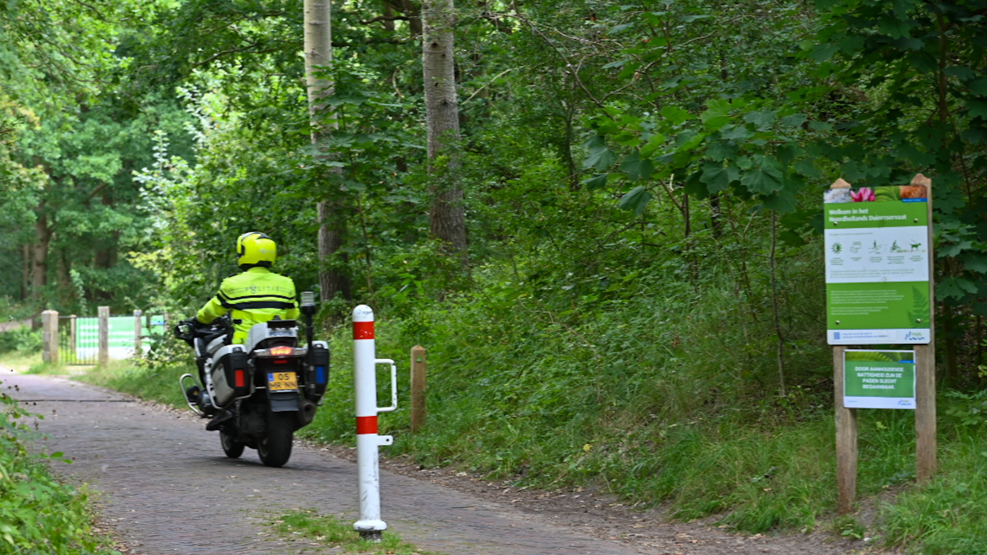 Politieagent op een motorfiets rijdt op een pad door een bos, naast een bord met informatie over het natuurgebied.