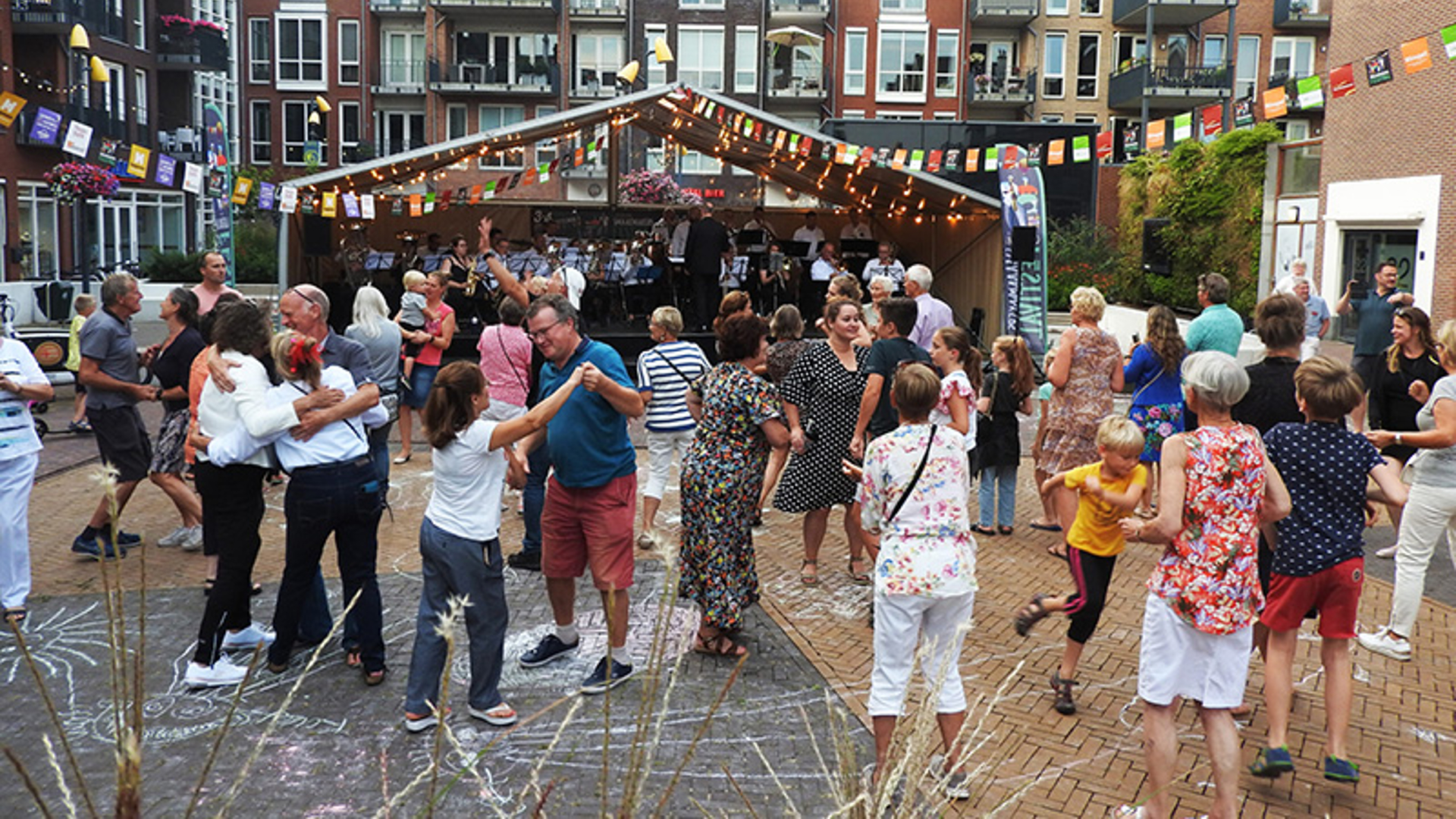 Mensen dansen en genieten van een buitenfestival met livemuziek onder een verlichte tent, omringd door feestelijke vlaggen en appartementsgebouwen.