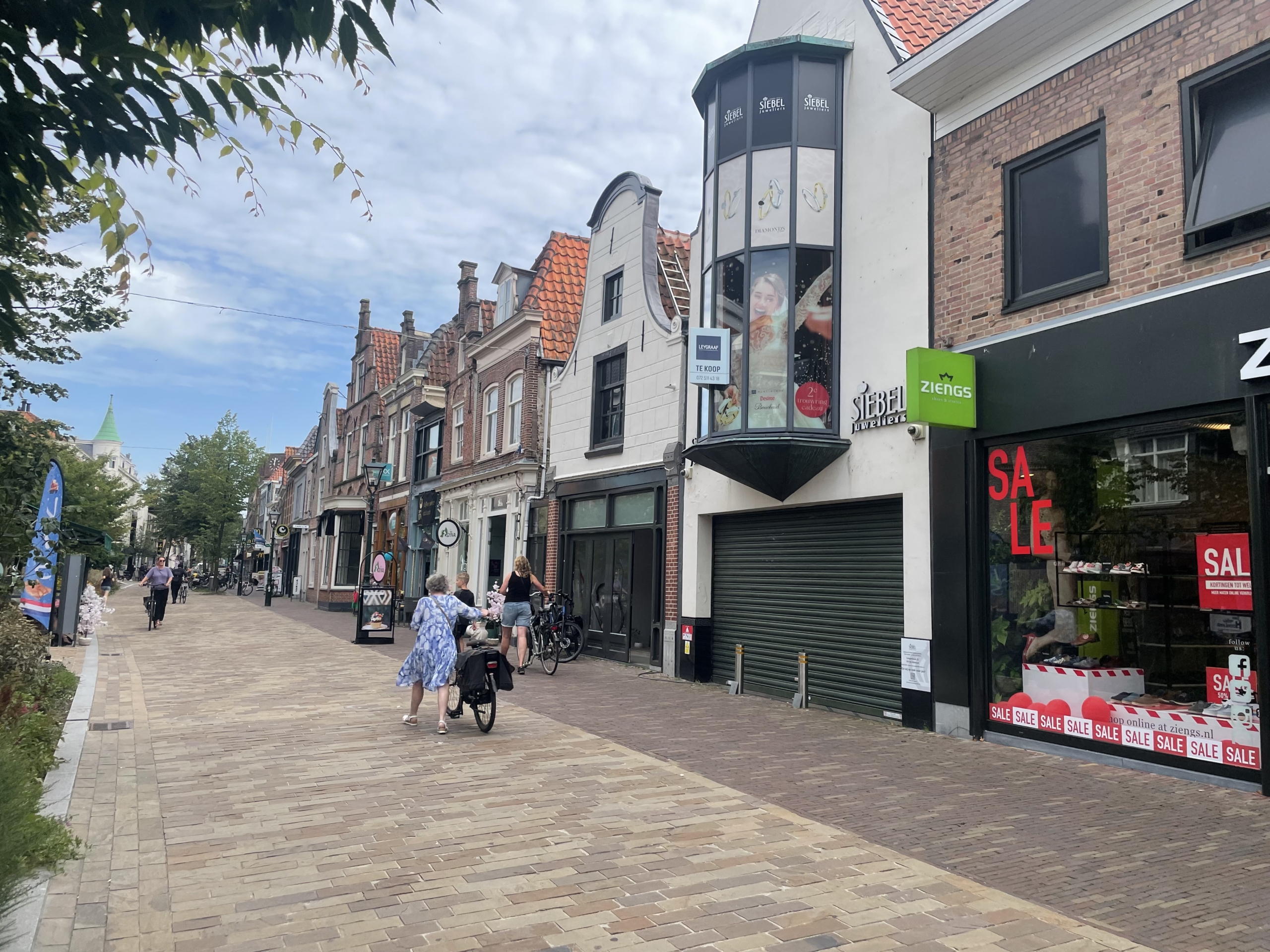 Winkelstraat met oude gebouwen en winkels, waaronder Ziengs en een gesloten Siebel juweliers, met wandelende mensen en fietsen.