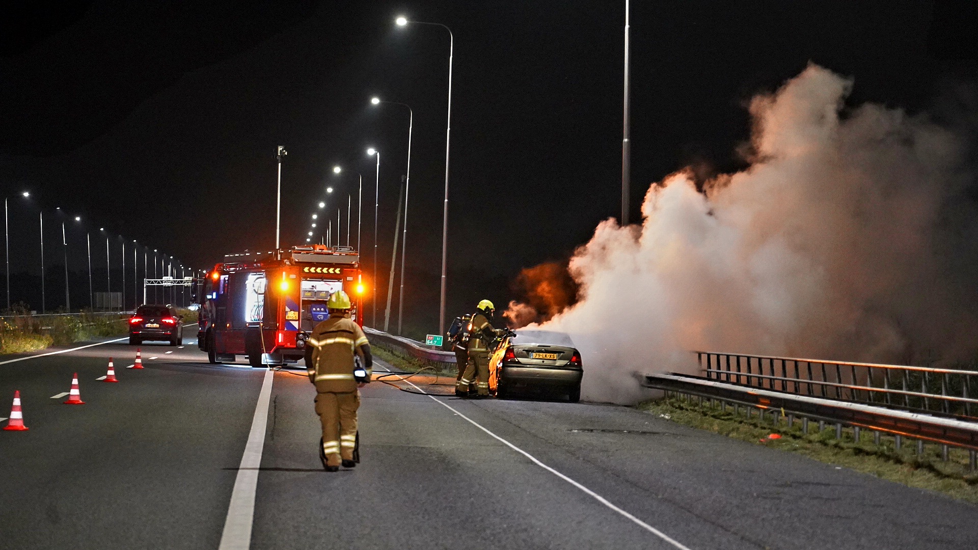 Brandweer blust brandende auto op snelweg 's nachts, met brandweerwagen en kegelmarkeringen op de achtergrond.